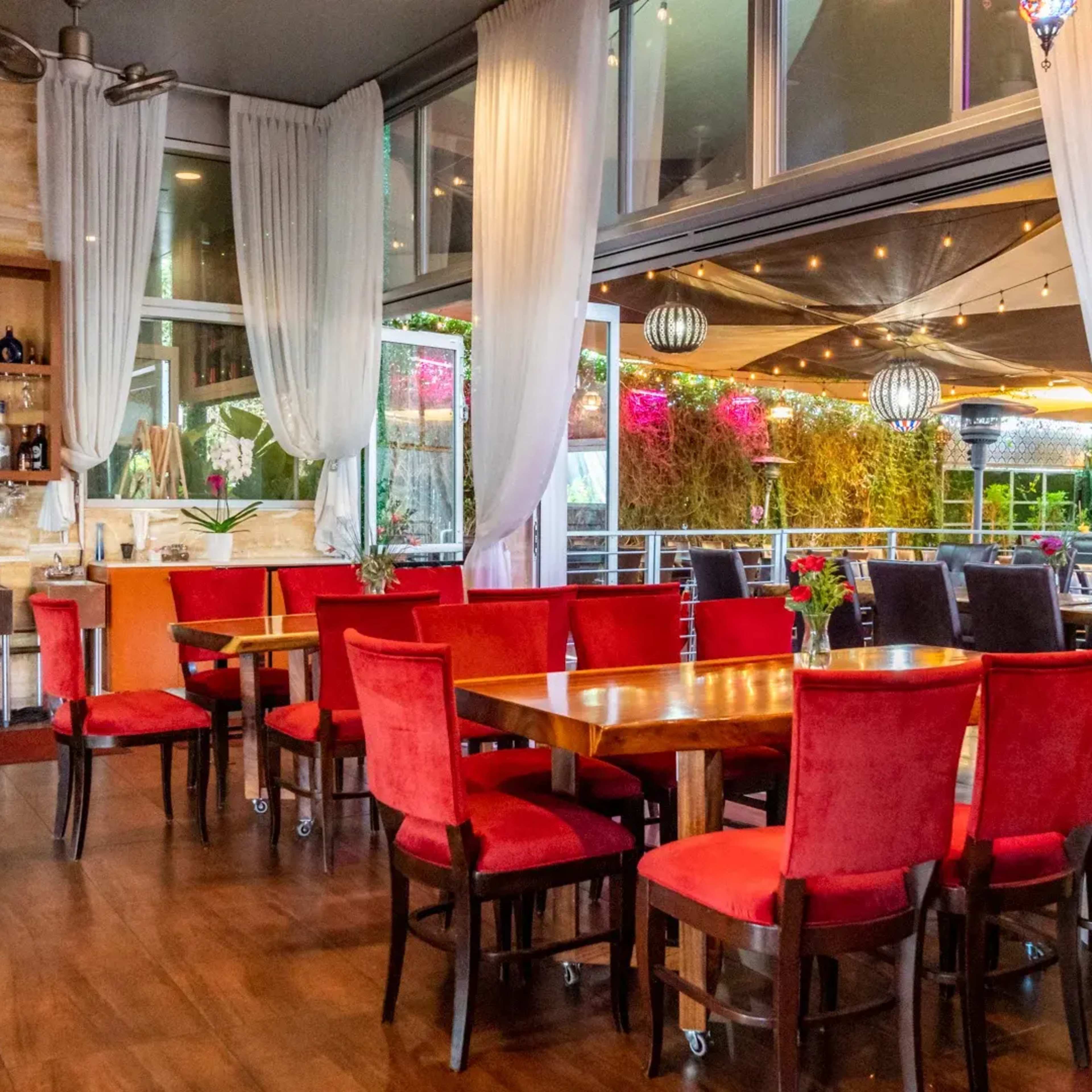 A restaurant interior with red upholstered chairs around wooden tables, large windows, and a bar area featuring shelves of bottles.