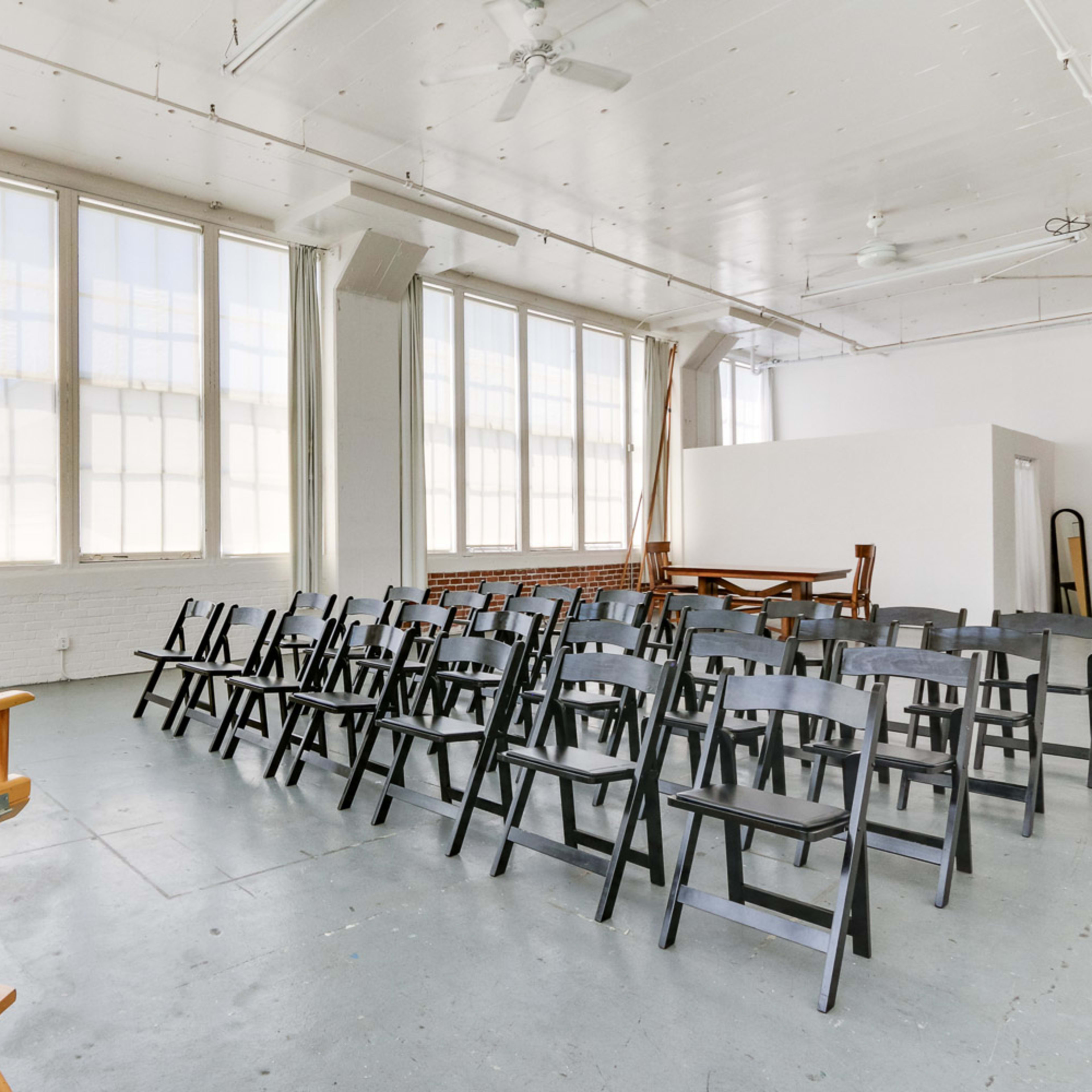 A bright, spacious room with large windows features rows of black chairs facing a wooden director's chair.