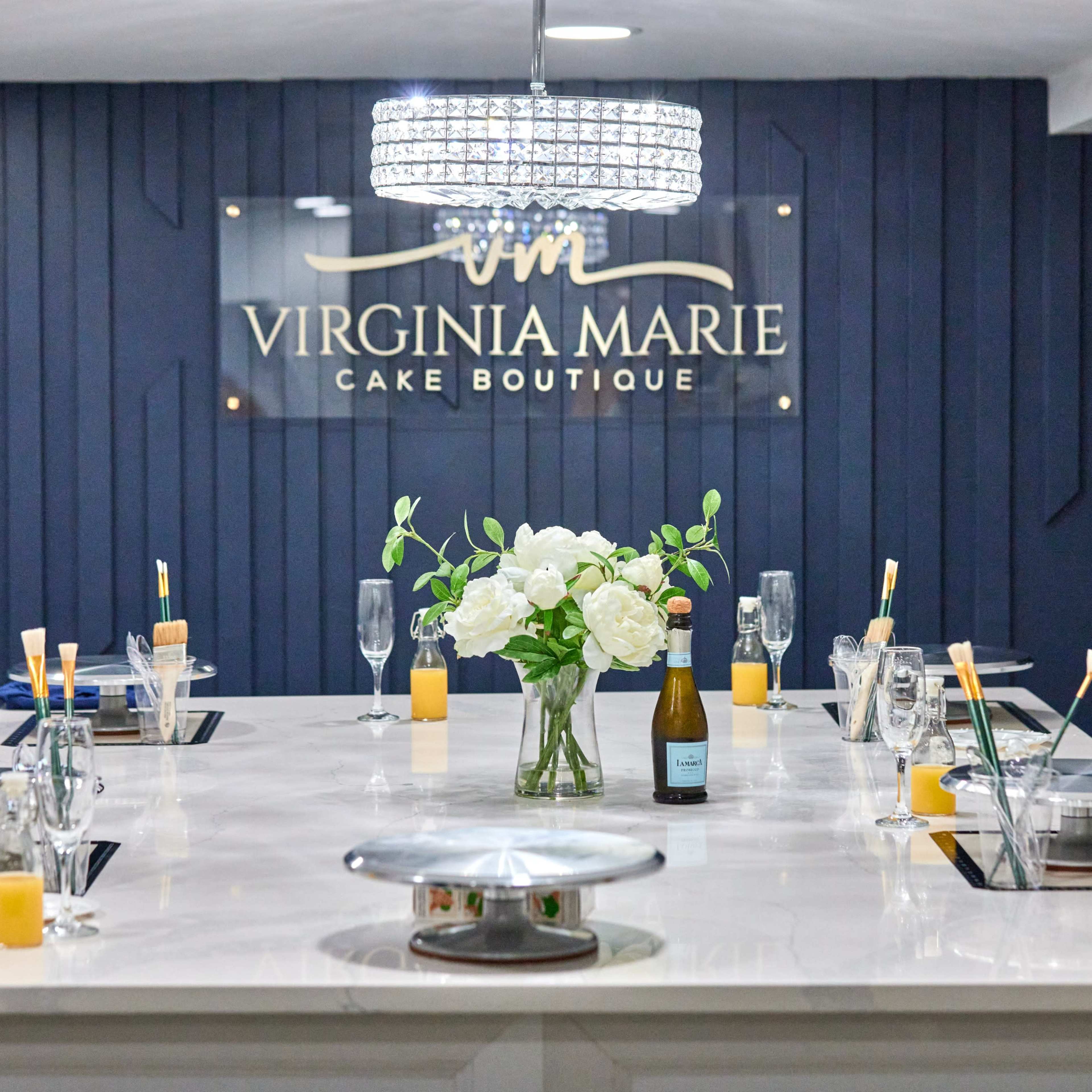 A modern cake boutique with a large, elegant table set for a gathering, featuring a floral centerpiece, sparkling chandelier, and branded wall sign.
