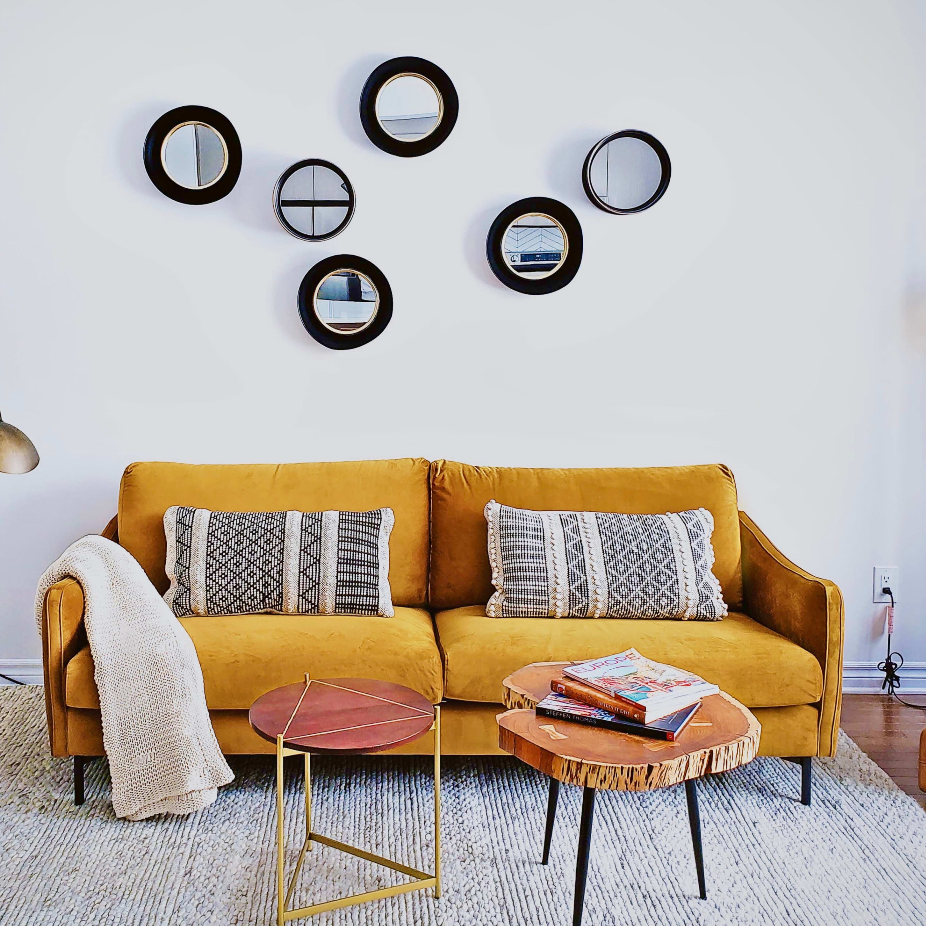 A cozy living room featuring a mustard yellow sofa with patterned cushions, surrounded by two side tables, a floor lamp, and a wall adorned with circular mirrors.