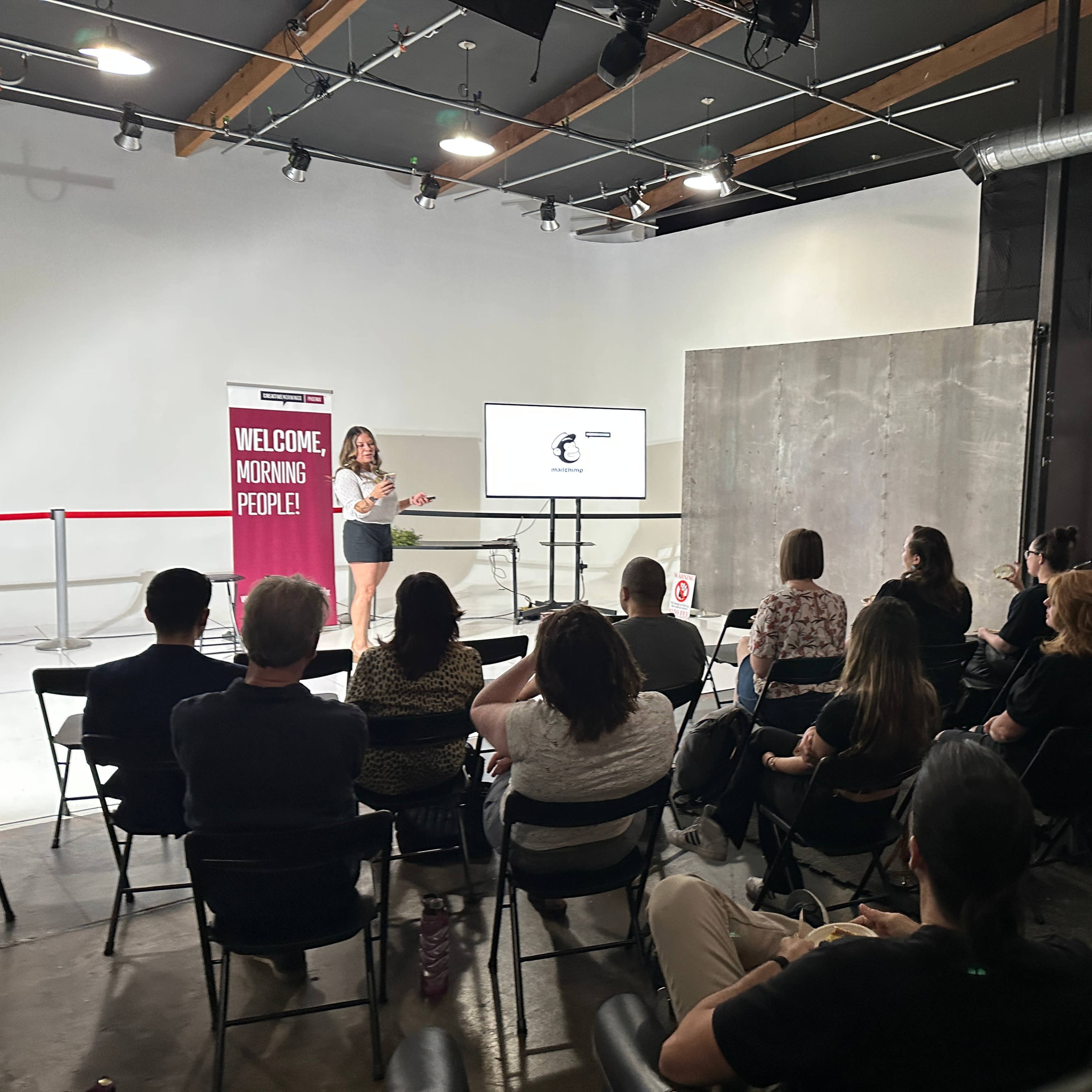 A speaker presents to an audience sitting in chairs at an event in a well-lit room with modern decor.