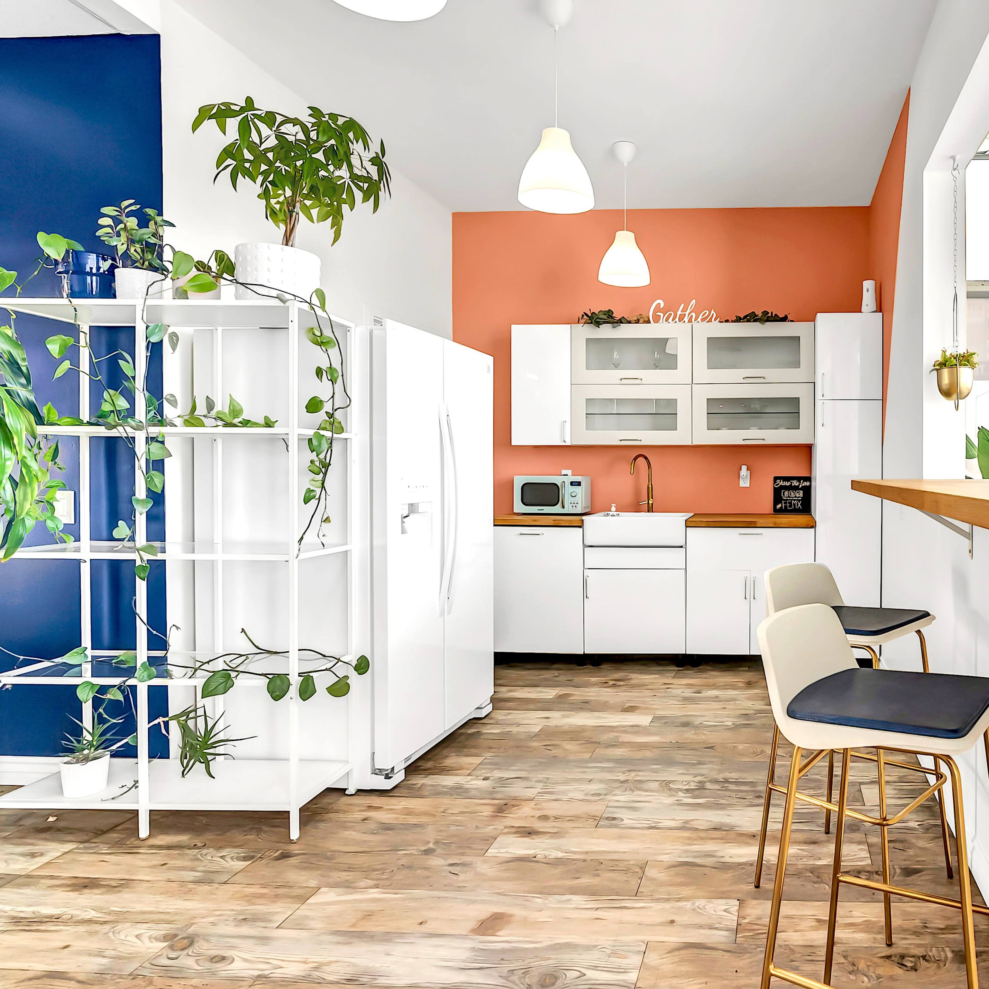 A modern kitchen with white cabinets, a blue accent wall, a wooden countertop with stools, and plants decorating the space.
