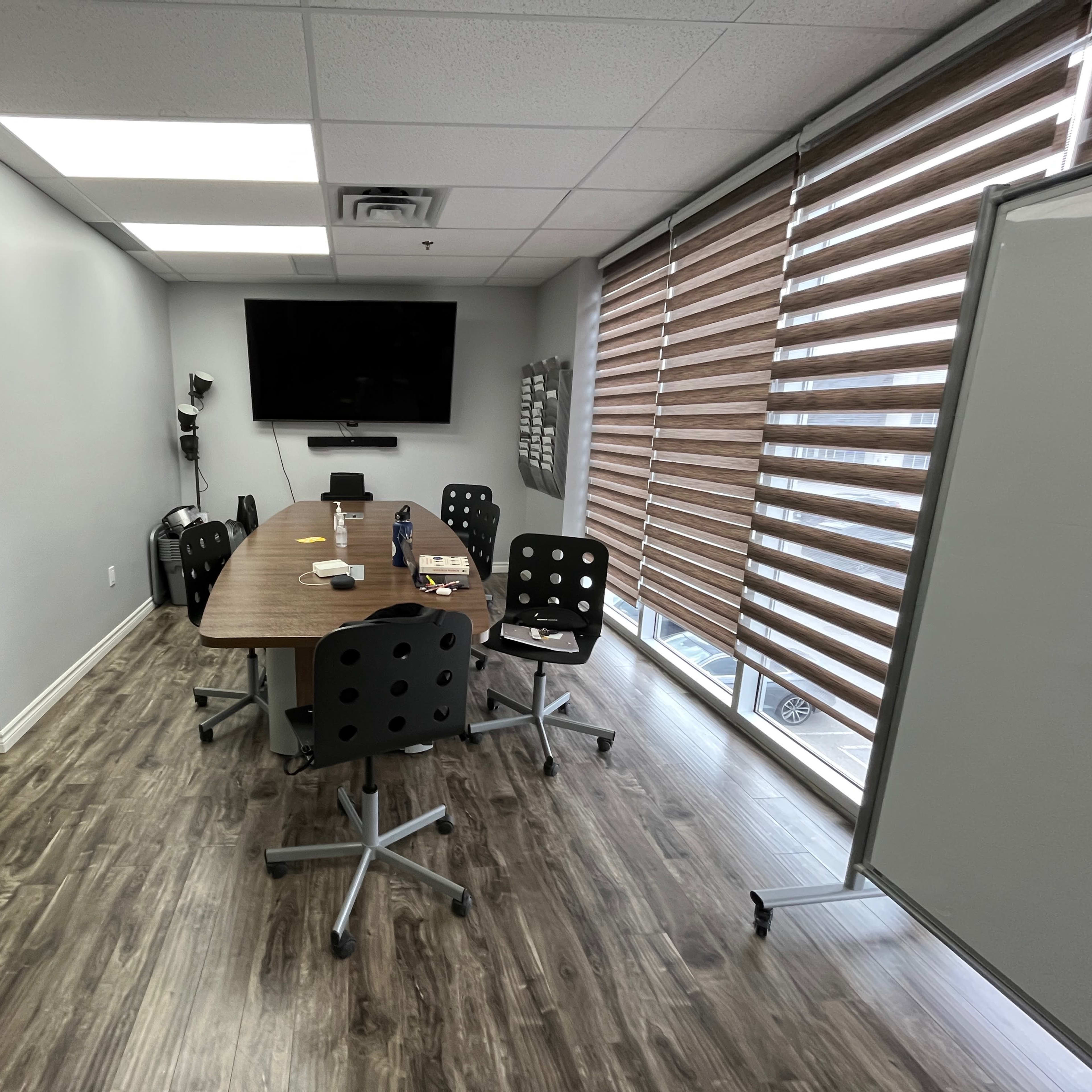 A modern conference room features a long wooden table surrounded by six rolling chairs, with a large screen mounted on the wall and window blinds partially closed.