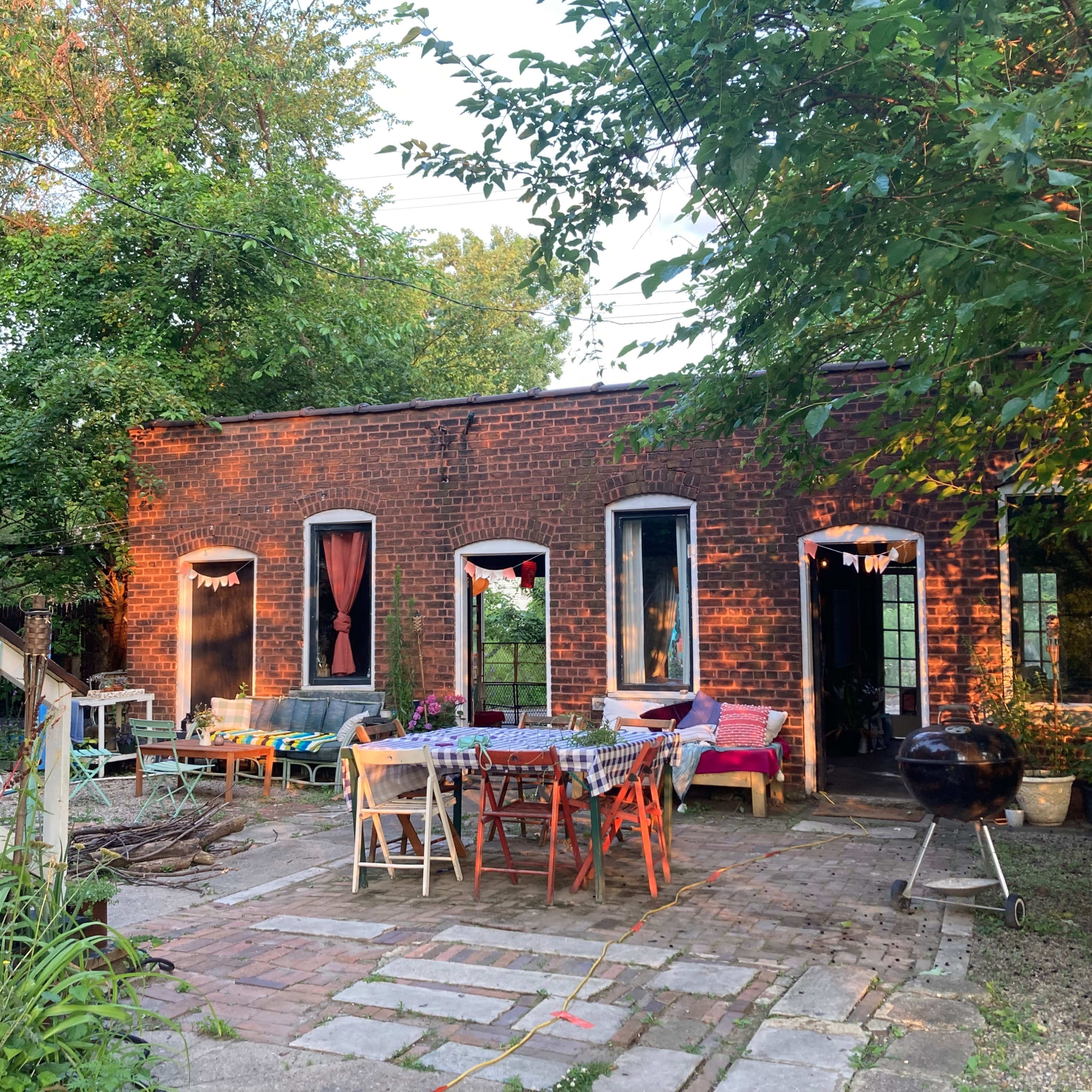 A brick building with multiple windows houses a patio area furnished with various tables and chairs, surrounded by greenery.
