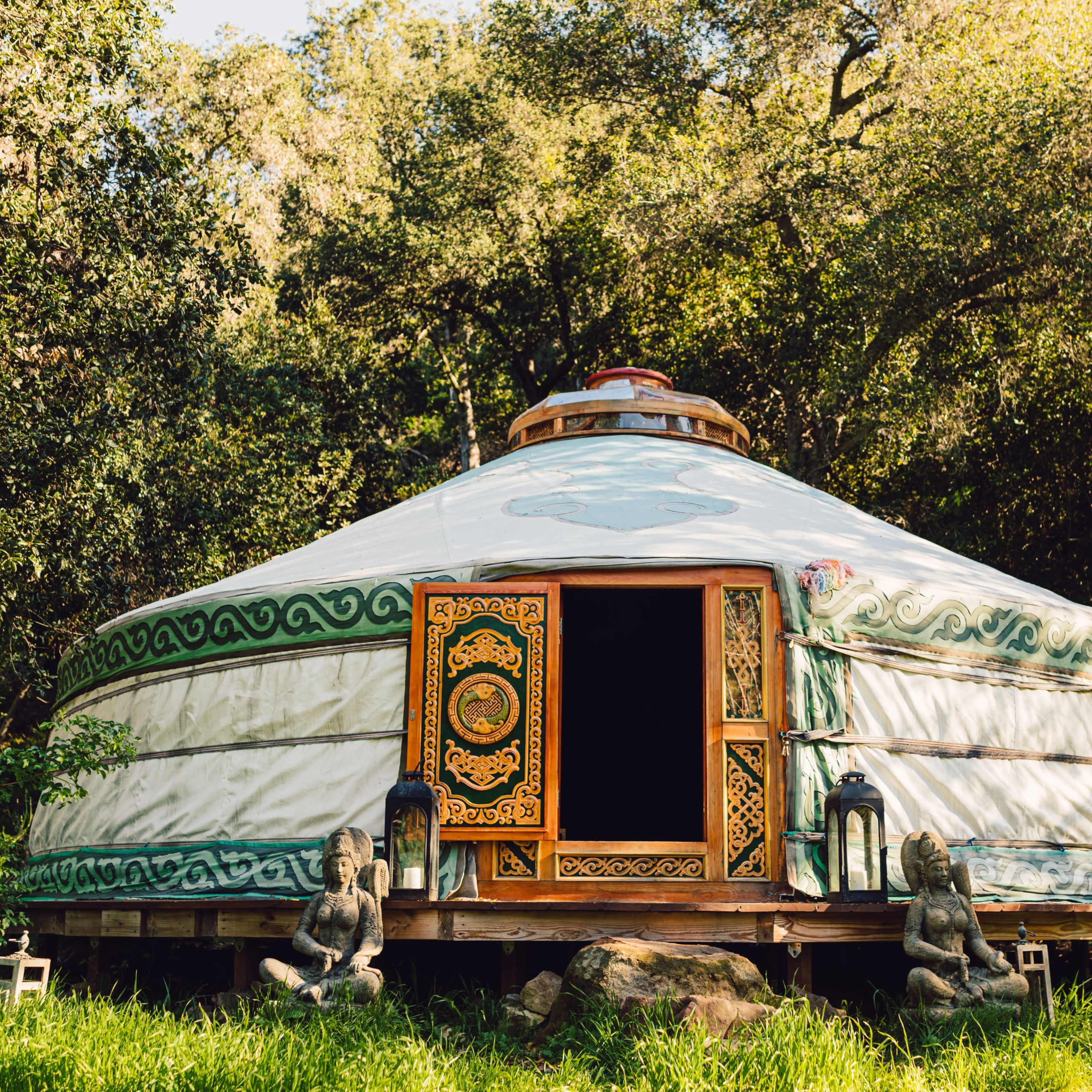 A traditional yurt with a wooden door and intricate designs is surrounded by greenery and features two stone statues in front.