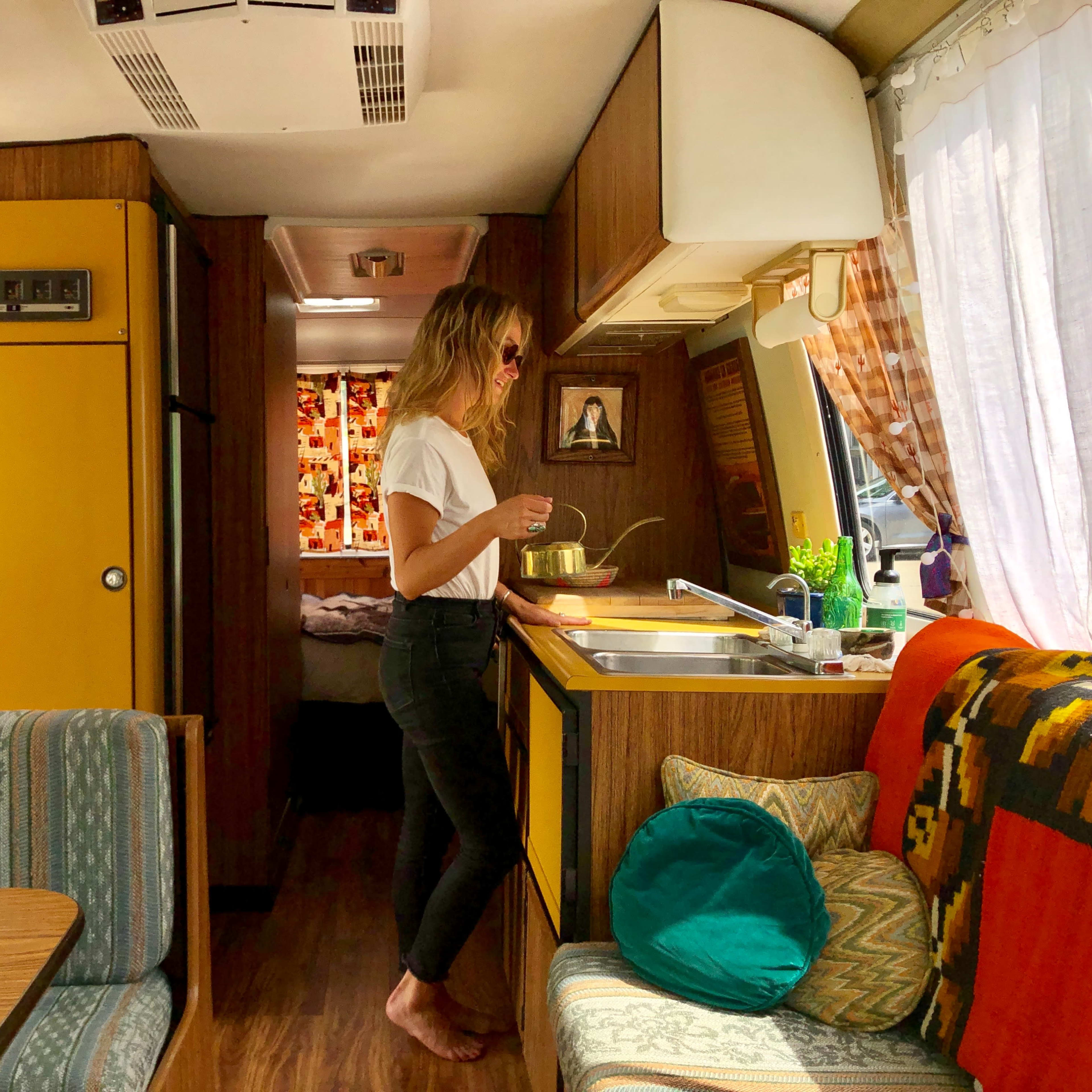 A woman stands in a retro-styled camper van, preparing food at a kitchen counter with vibrant decor.