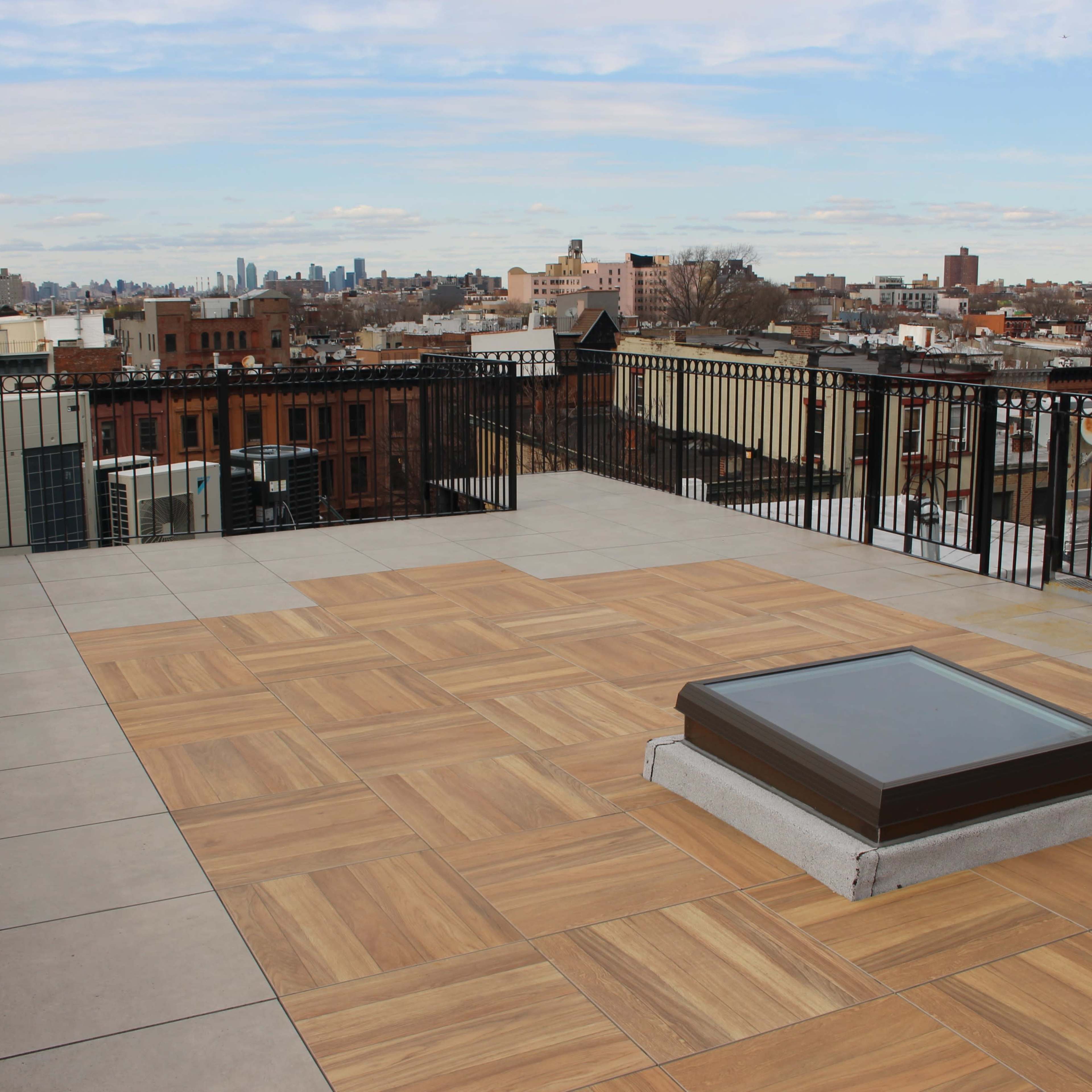 A spacious rooftop terrace with a wooden-patterned floor and a skylight, overlooking a city skyline in the background.