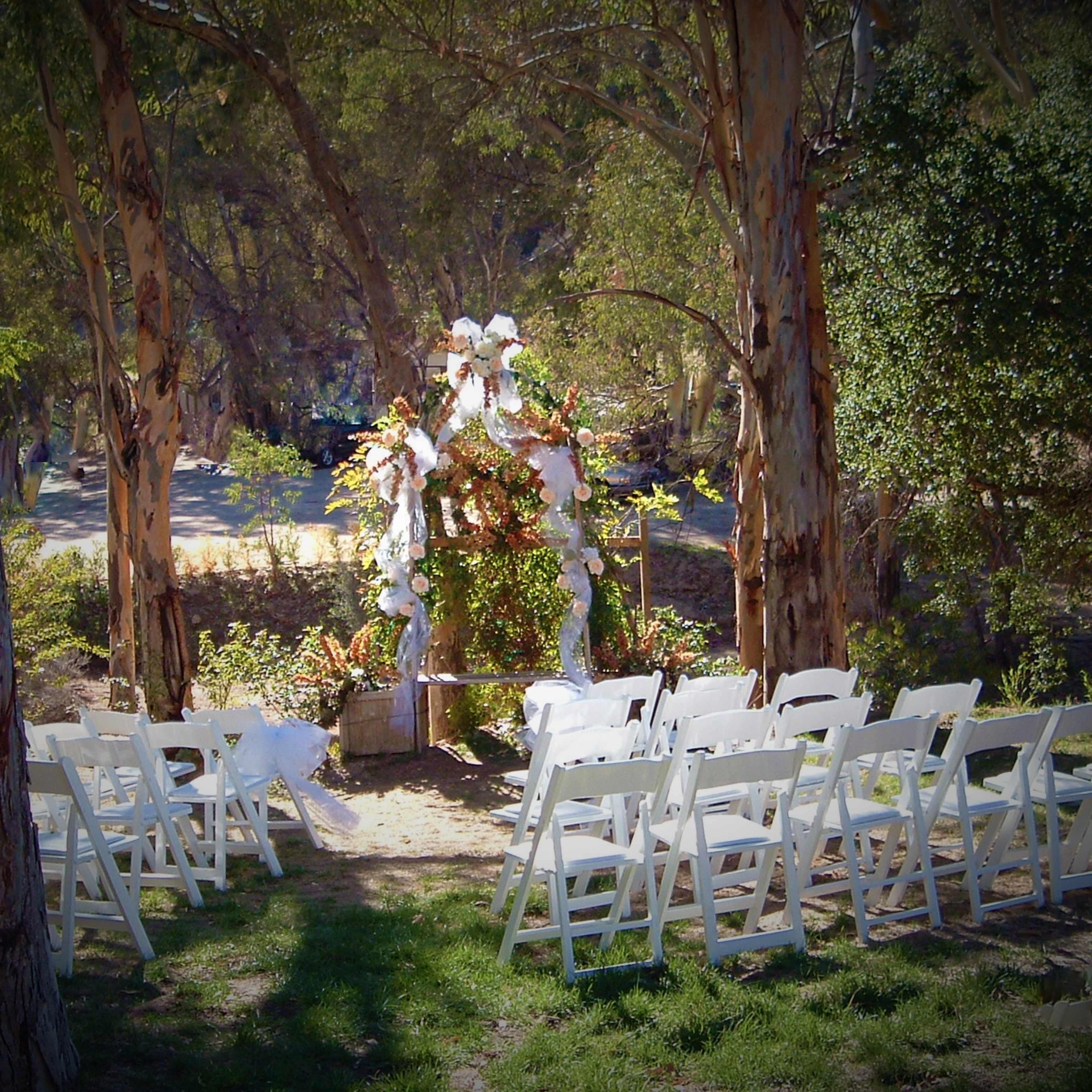 A decorated outdoor wedding altar is set up among trees, facing rows of white chairs arranged for guests.