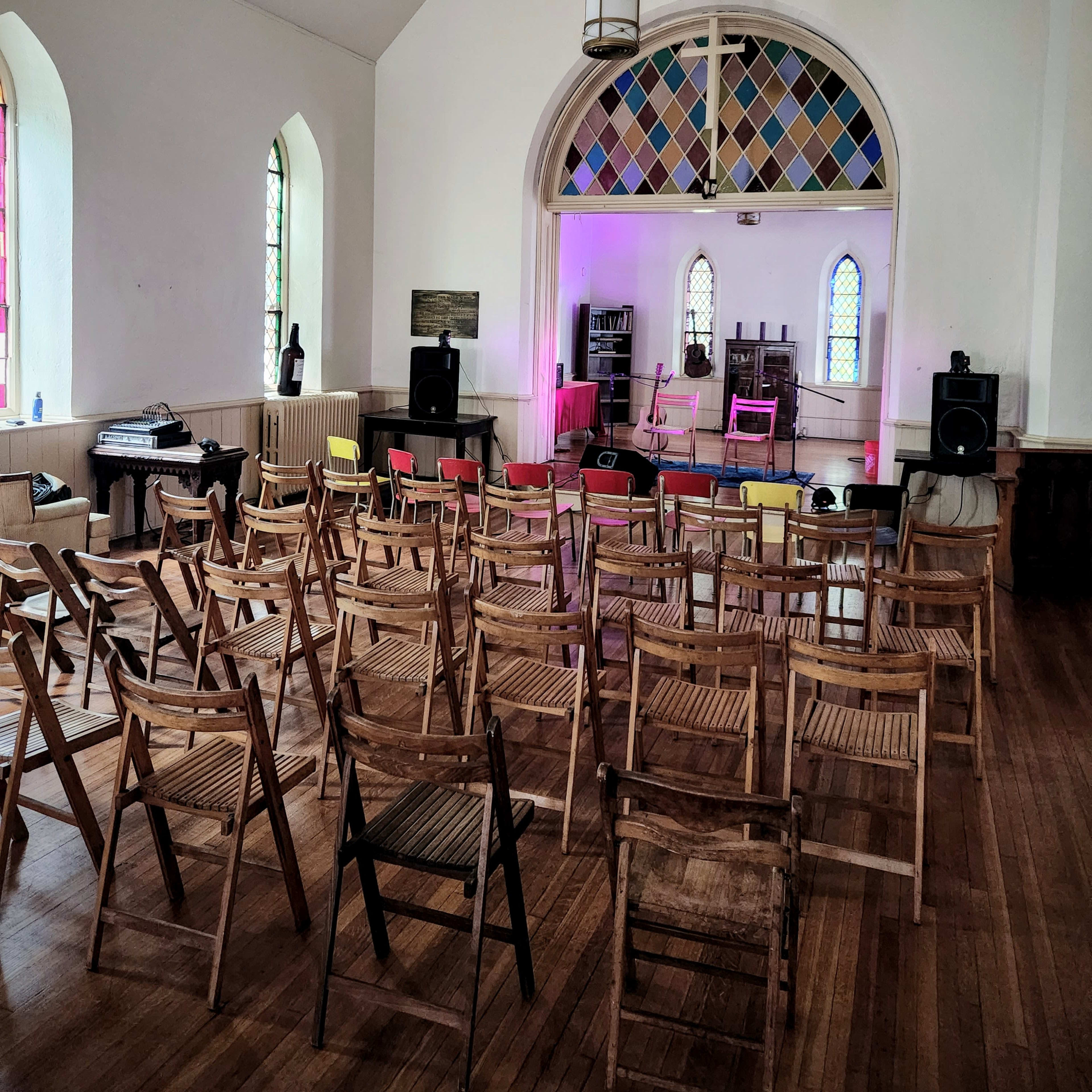 A sparsely decorated room with folding chairs arranged in rows, a small stage at the front, and colorful lights illuminating the space.