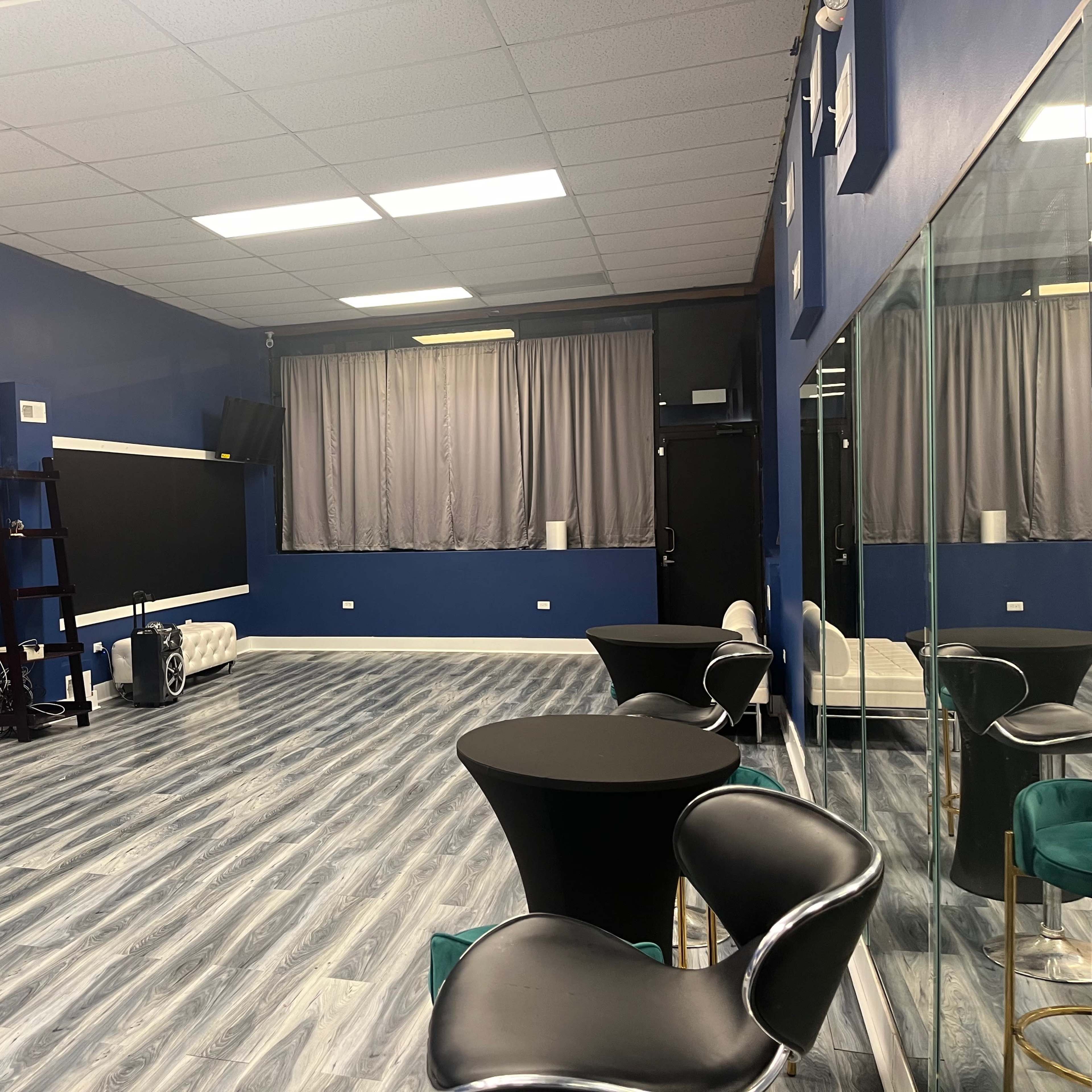 A spacious, empty room with blue walls, gray wooden flooring, a large mirror along one wall, and two black tables with chairs.