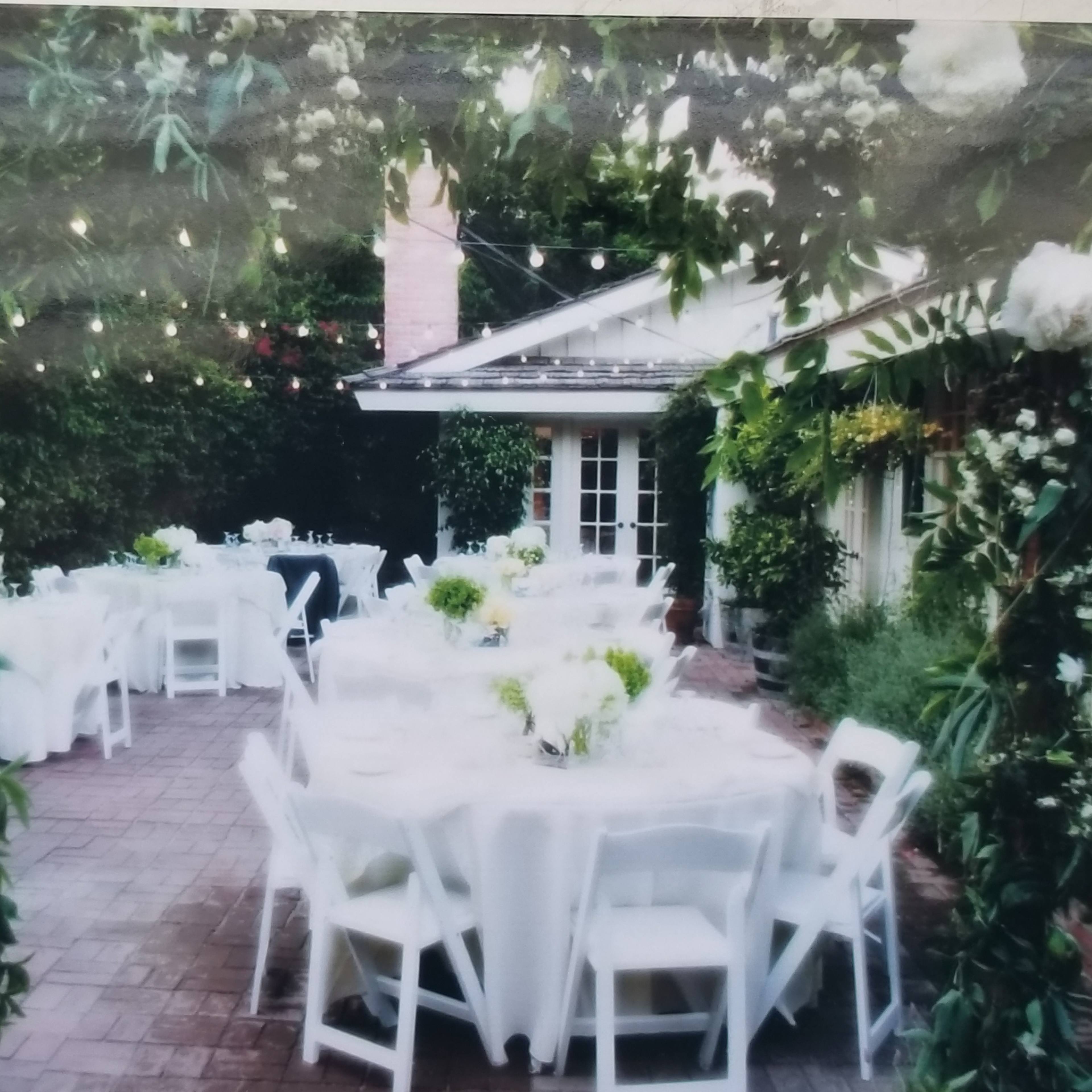 A beautifully arranged outdoor dining area with white tables and chairs surrounded by greenery and string lights.