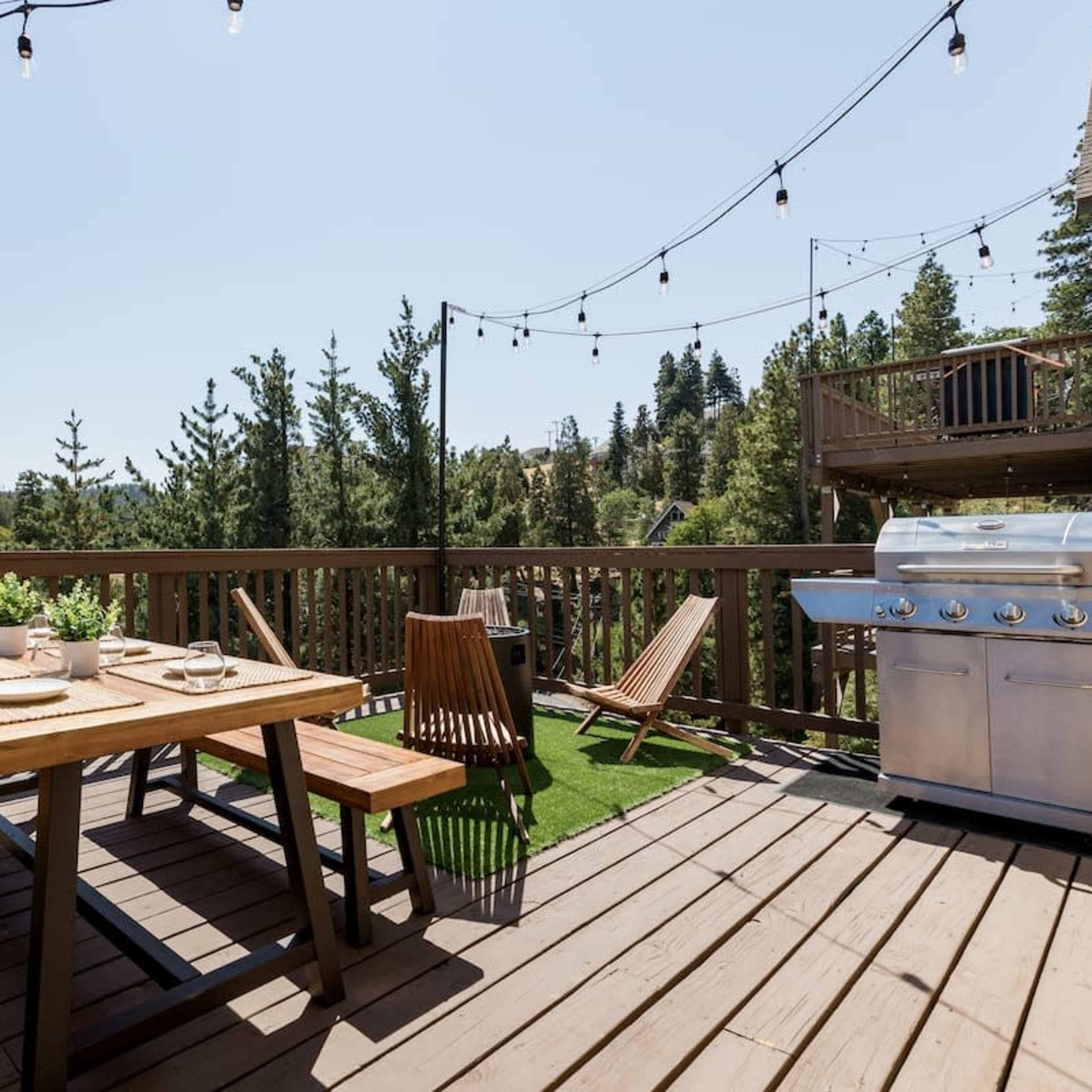 A wooden deck features a picnic table set with dishes and a gas grill, surrounded by tall trees and string lights overhead.