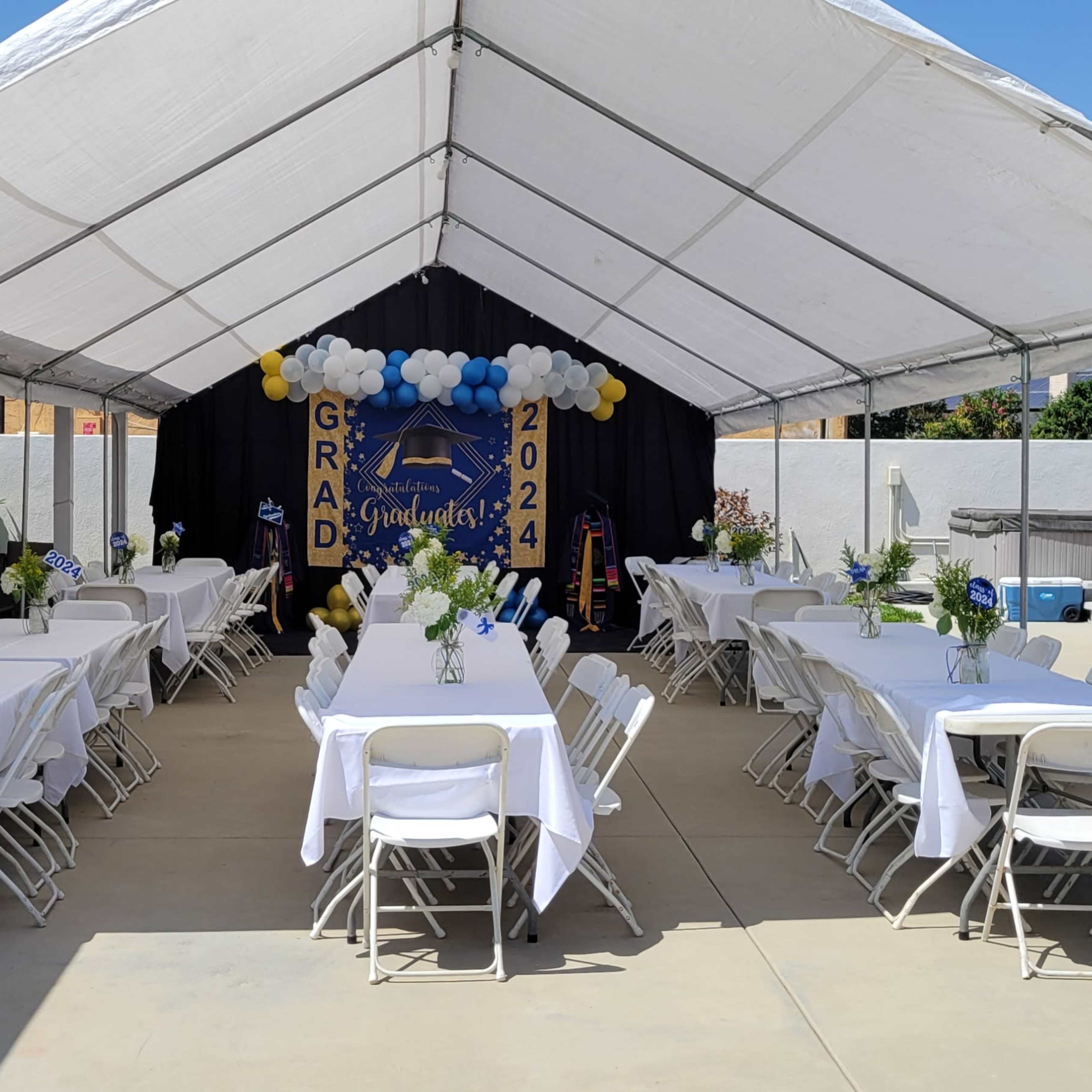A tented outdoor space is set up with white tablecloths and centerpieces for a graduation celebration.
