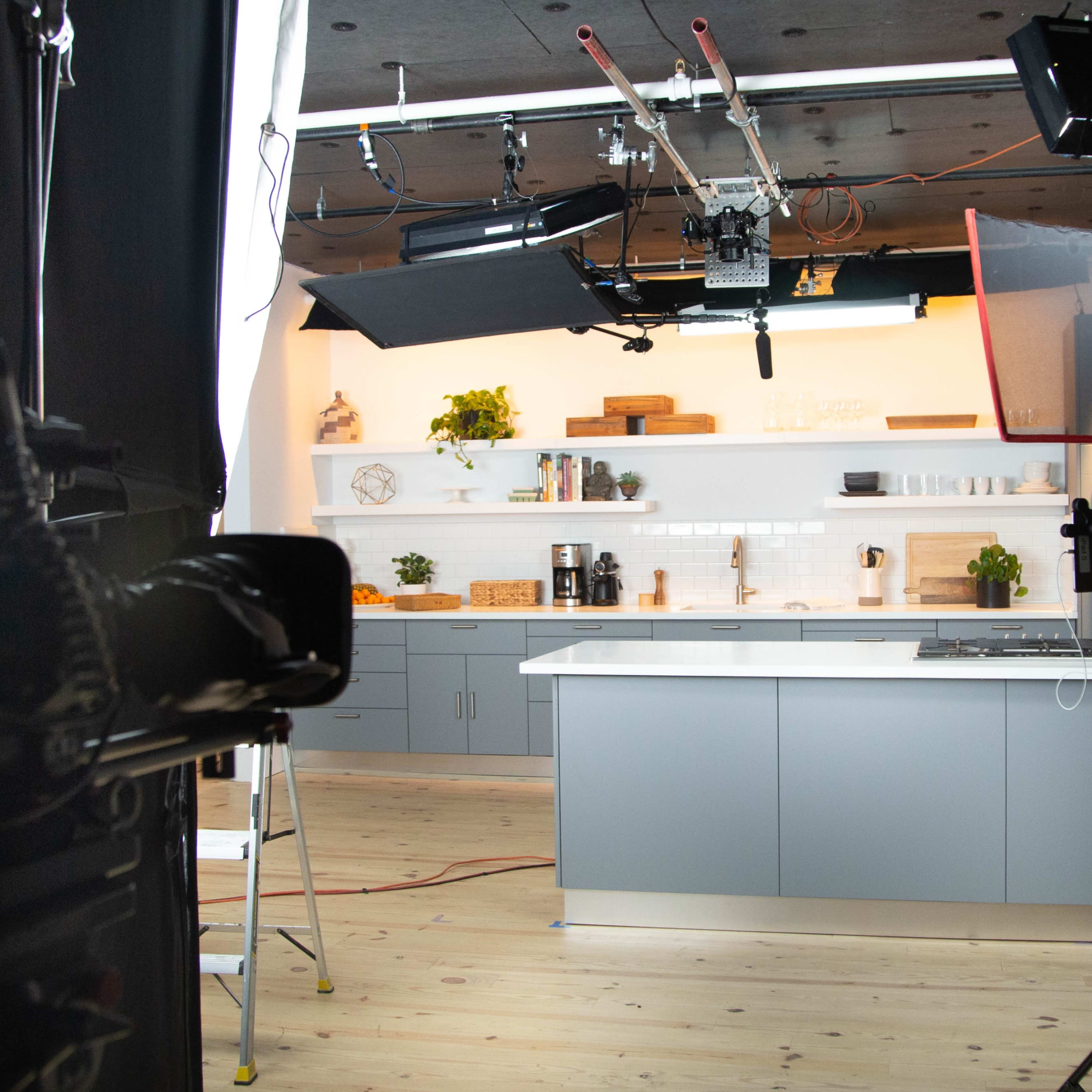 A well-equipped kitchen studio with a large gray island, various kitchen appliances, and multiple lights set up for filming.