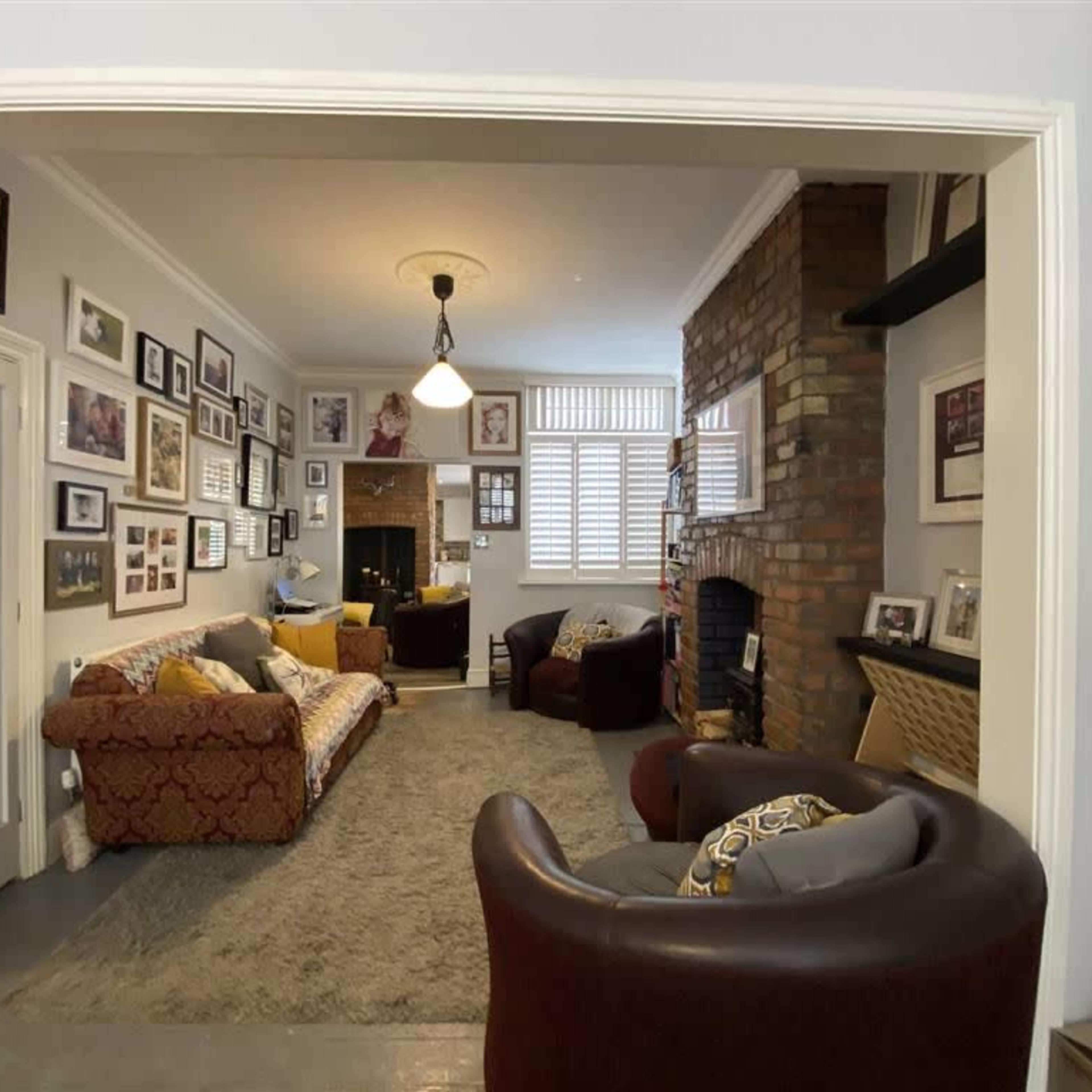 A cozy living room featuring a brick fireplace, framed photographs on the walls, and seating arrangements with a sofa and armchairs.
