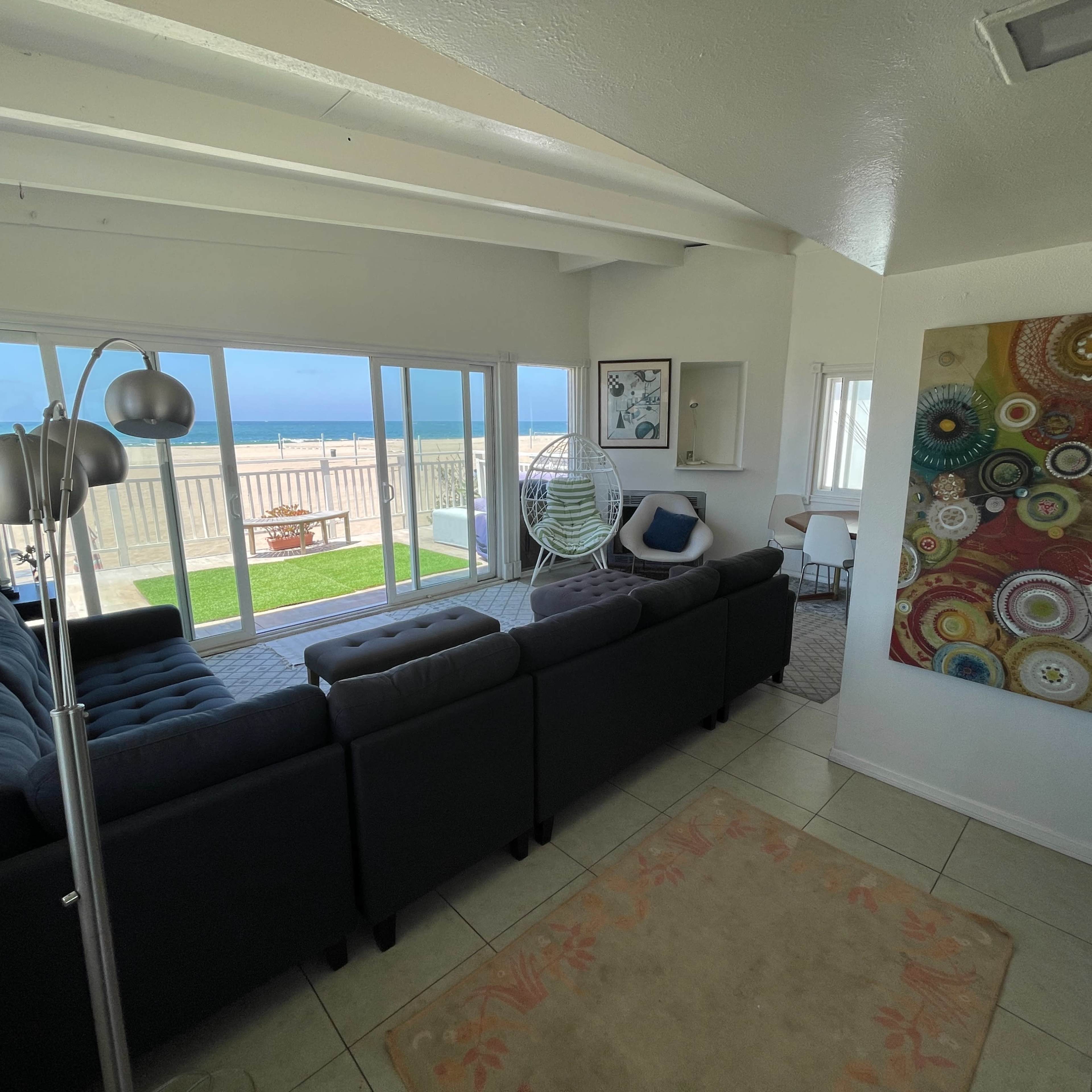 A modern living room features a dark sectional sofa, a colorful wall art piece, and large windows overlooking a grassy outdoor space.