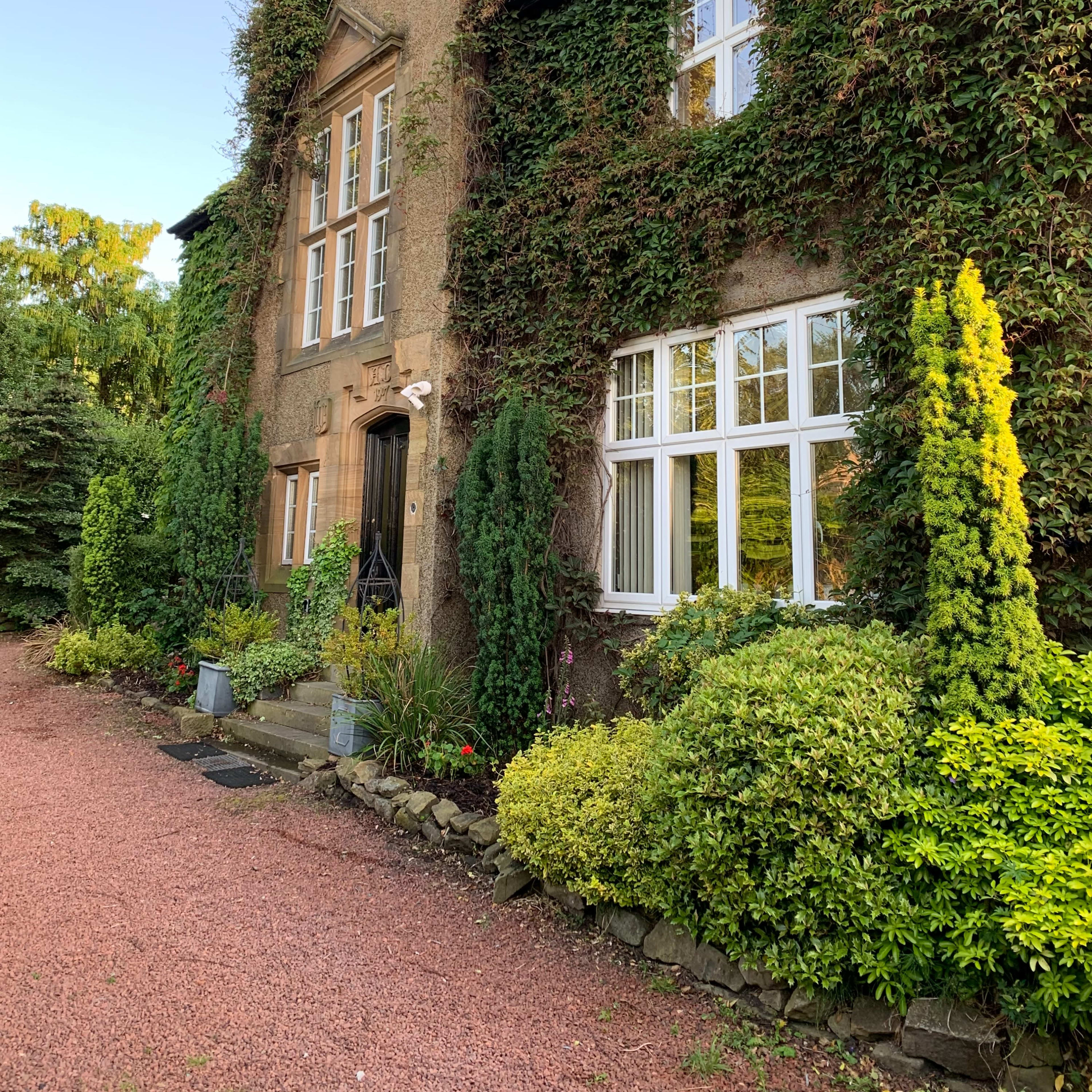 A two-story house adorned with ivy and surrounded by neatly arranged shrubs and flowers along a gravel pathway.