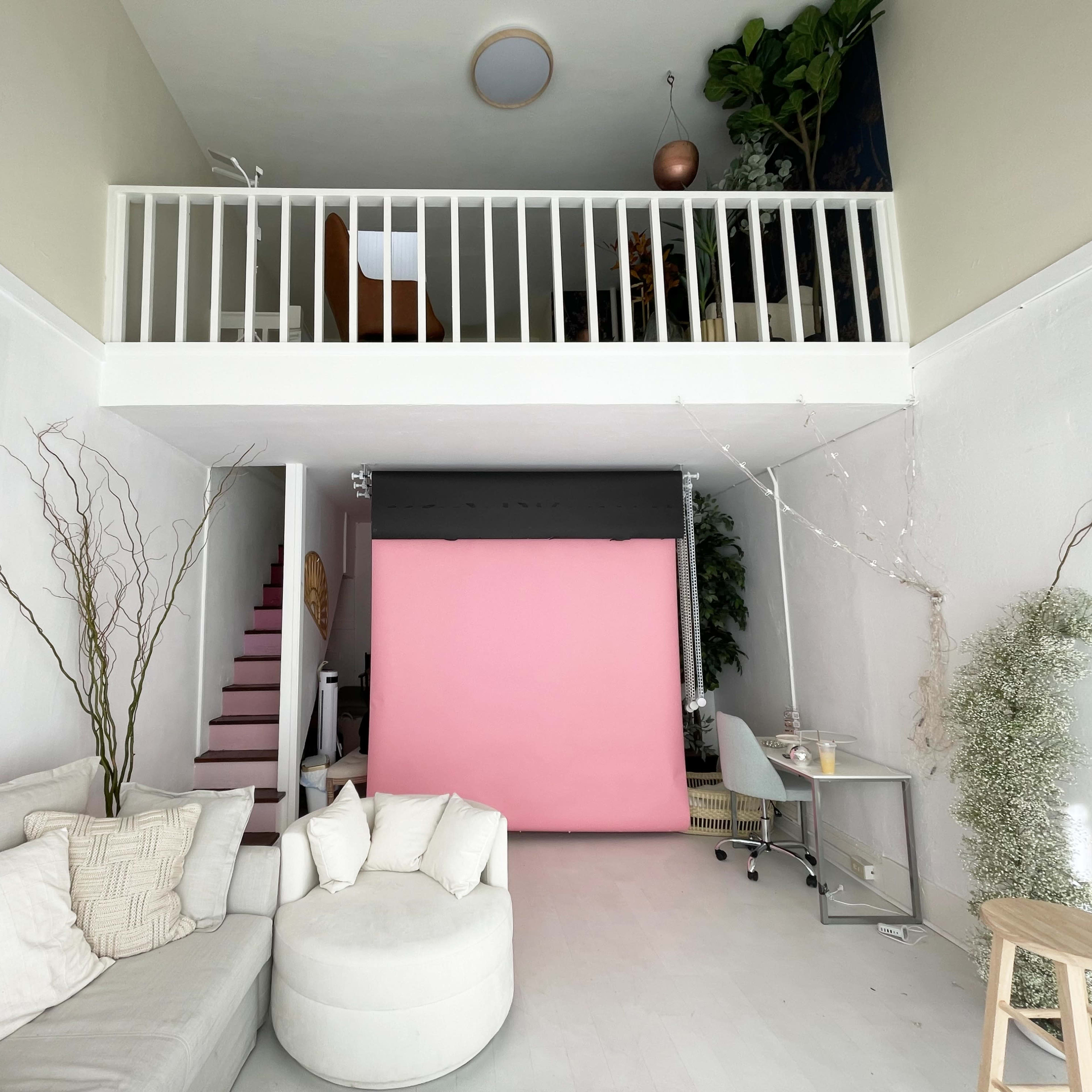 A spacious interior with a loft area, featuring a pink backdrop, a desk, a light-colored sofa, and decorative plants.