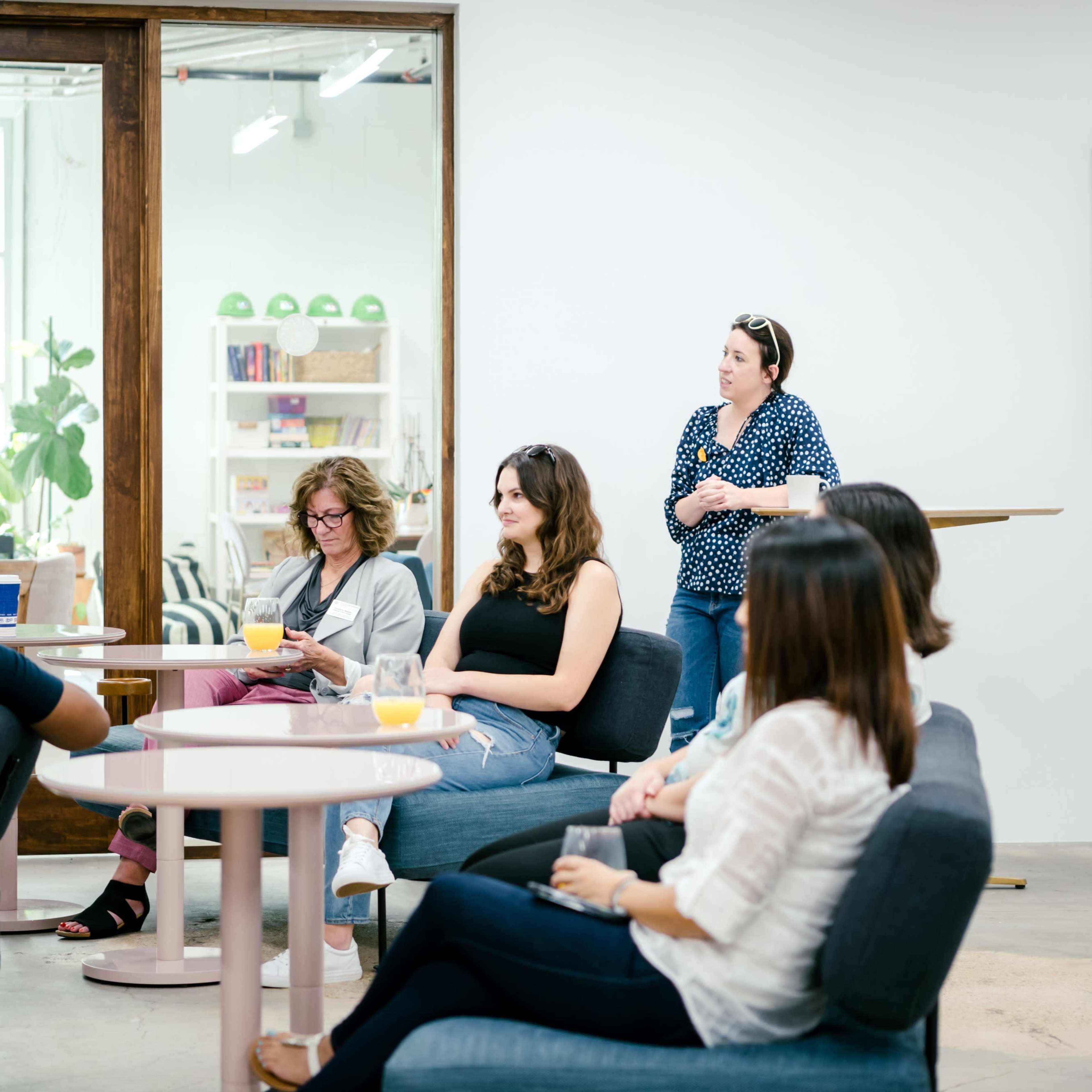 A group of women gathers in a modern, well-lit room with a casual seating arrangement, engaging in conversation.