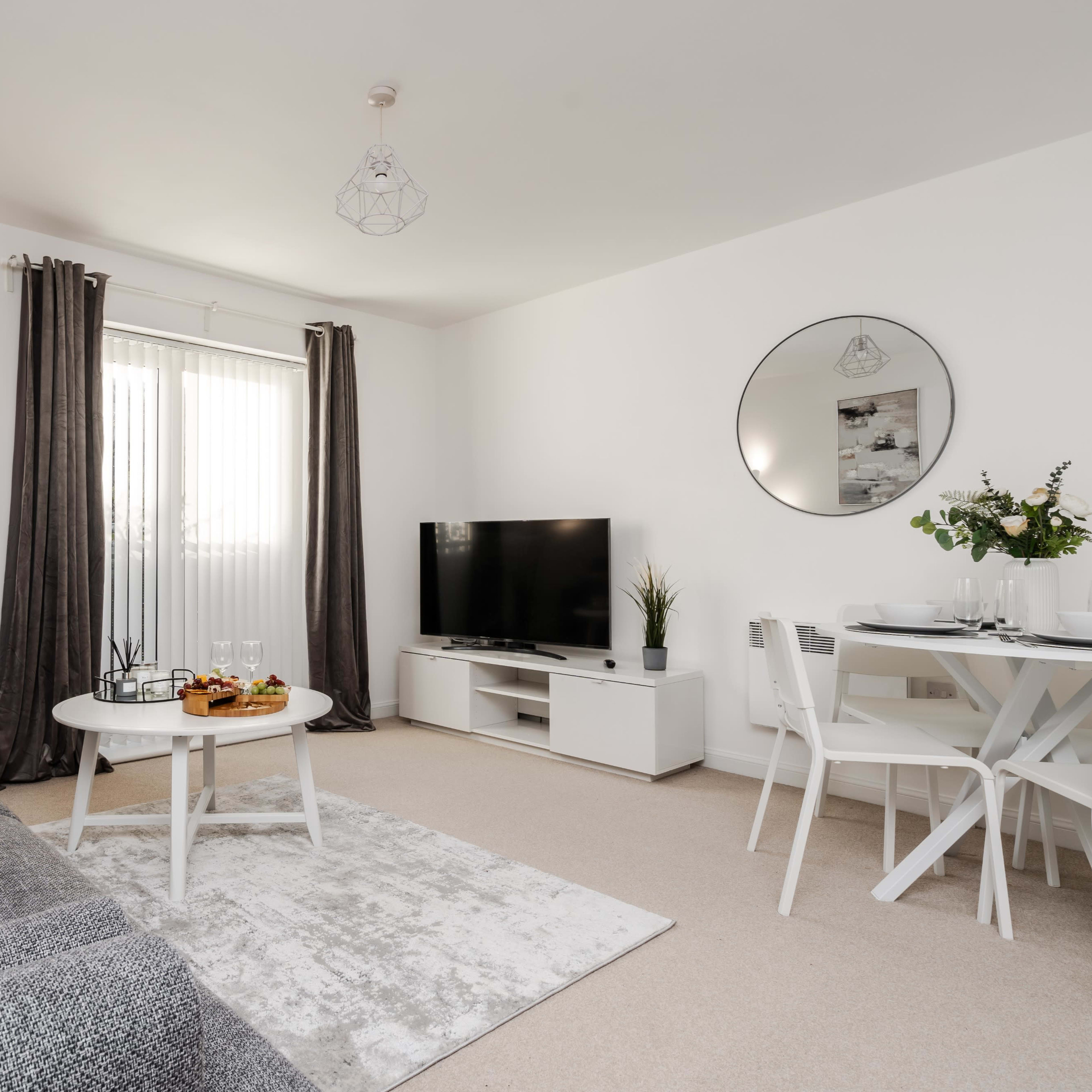 A modern living room features a gray sofa, a round coffee table with snacks, a flat-screen TV on a white unit, and a dining table set for four, all illuminated by natural light through sheer curtains.