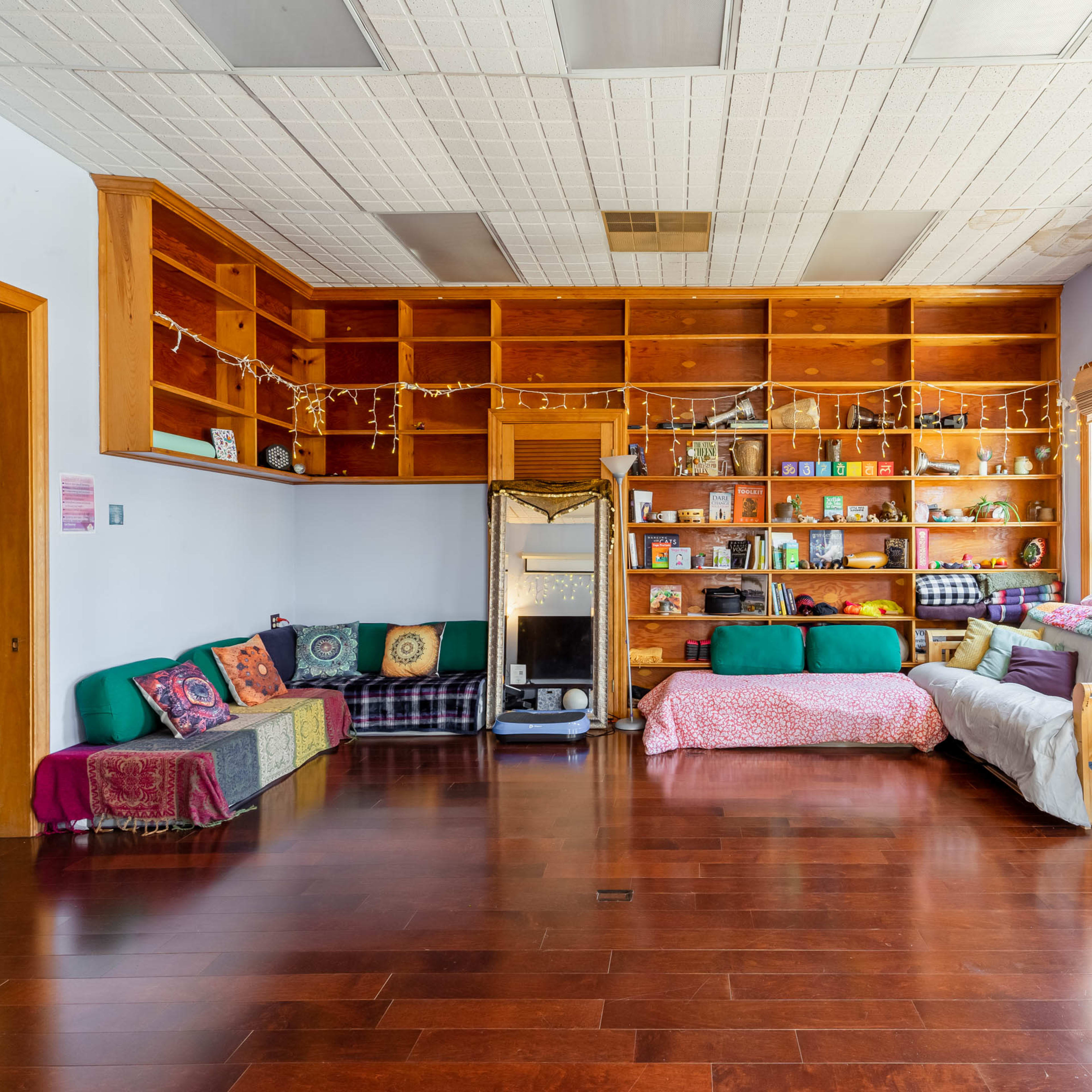 A spacious interior with a wooden floor, a wall of bookshelves filled with various items, and several couches and pillows arranged for seating.