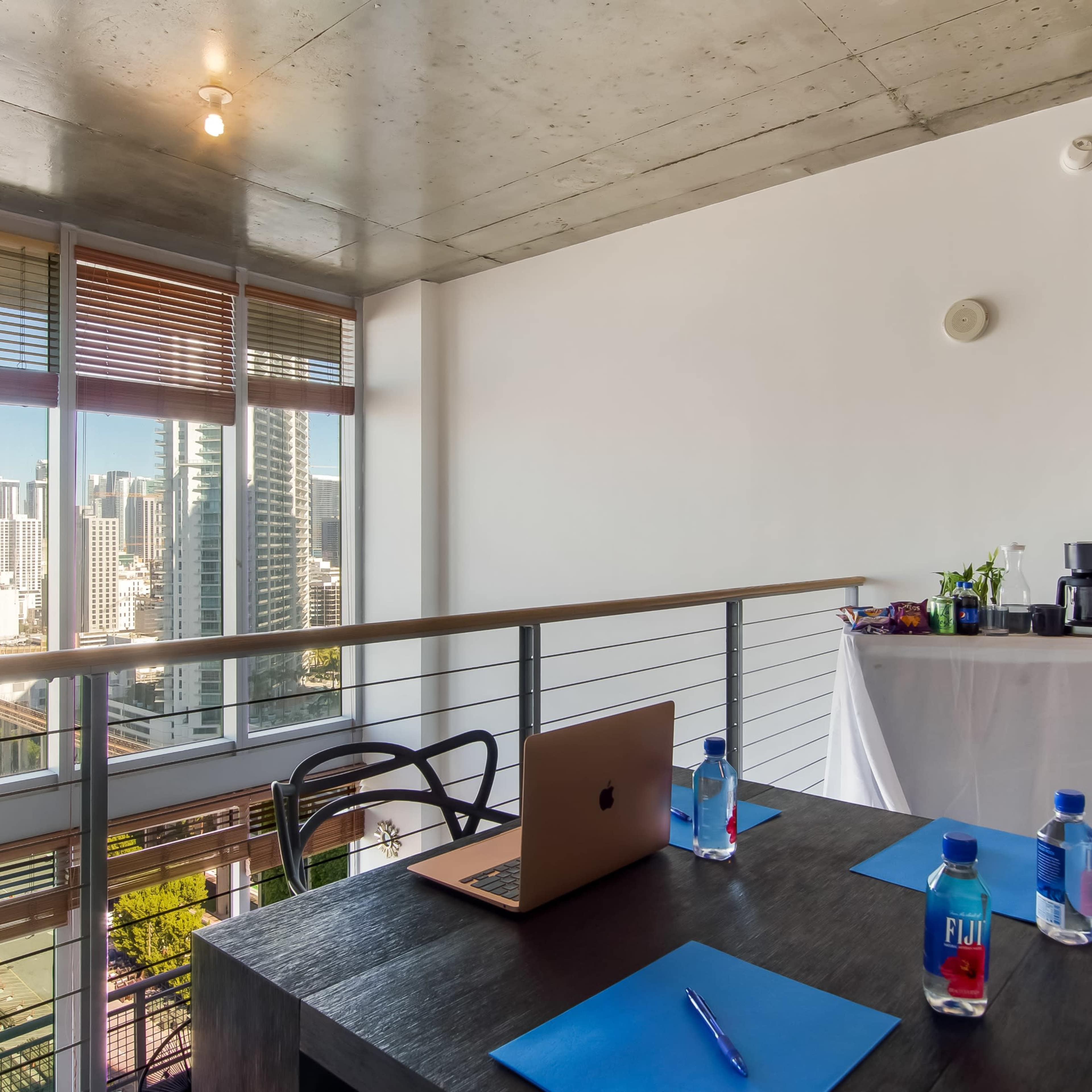 A modern workspace features a table with a laptop and water bottles, set against a backdrop of large windows overlooking a city skyline.