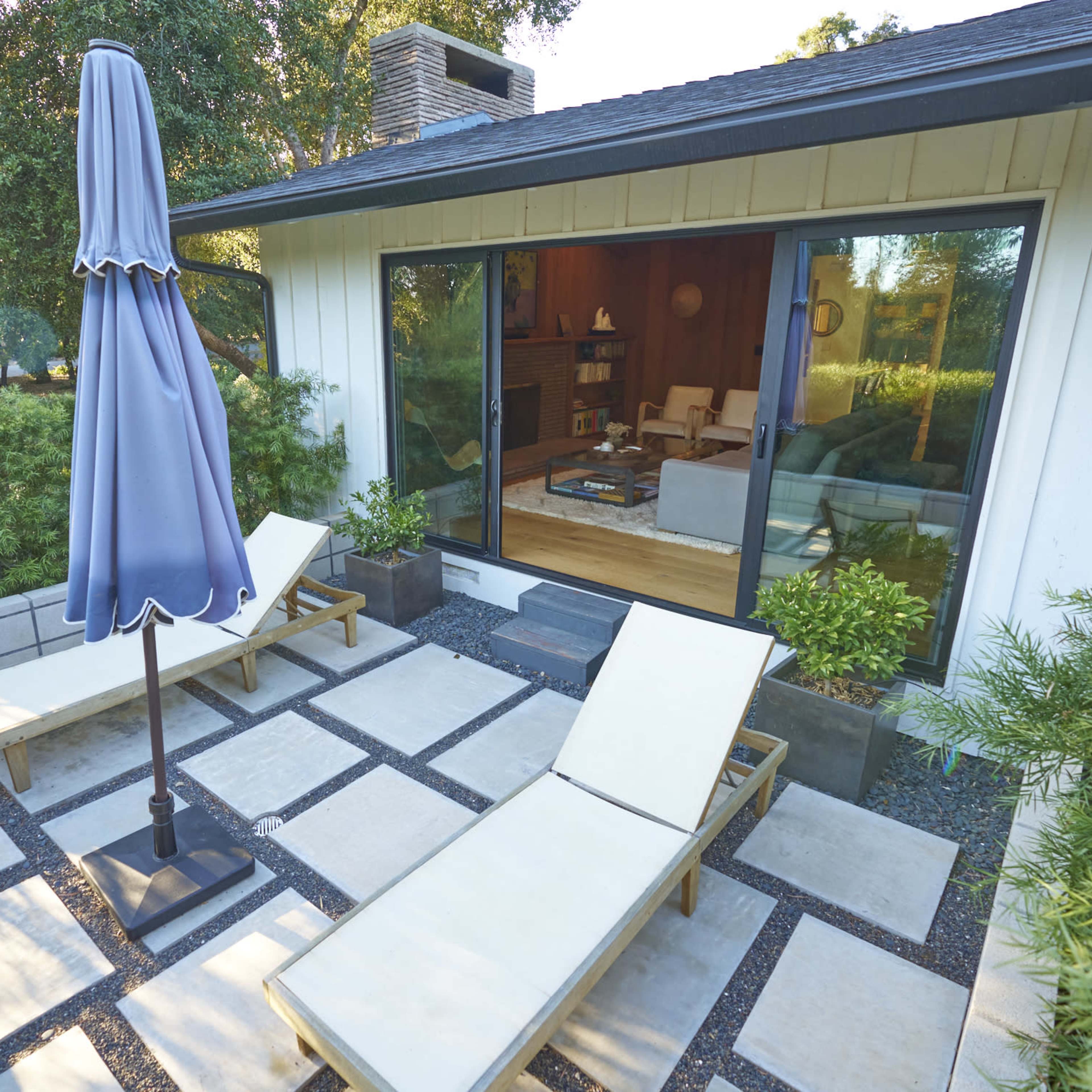 A patio with two lounge chairs and a large umbrella beside a modern house that features sliding glass doors.