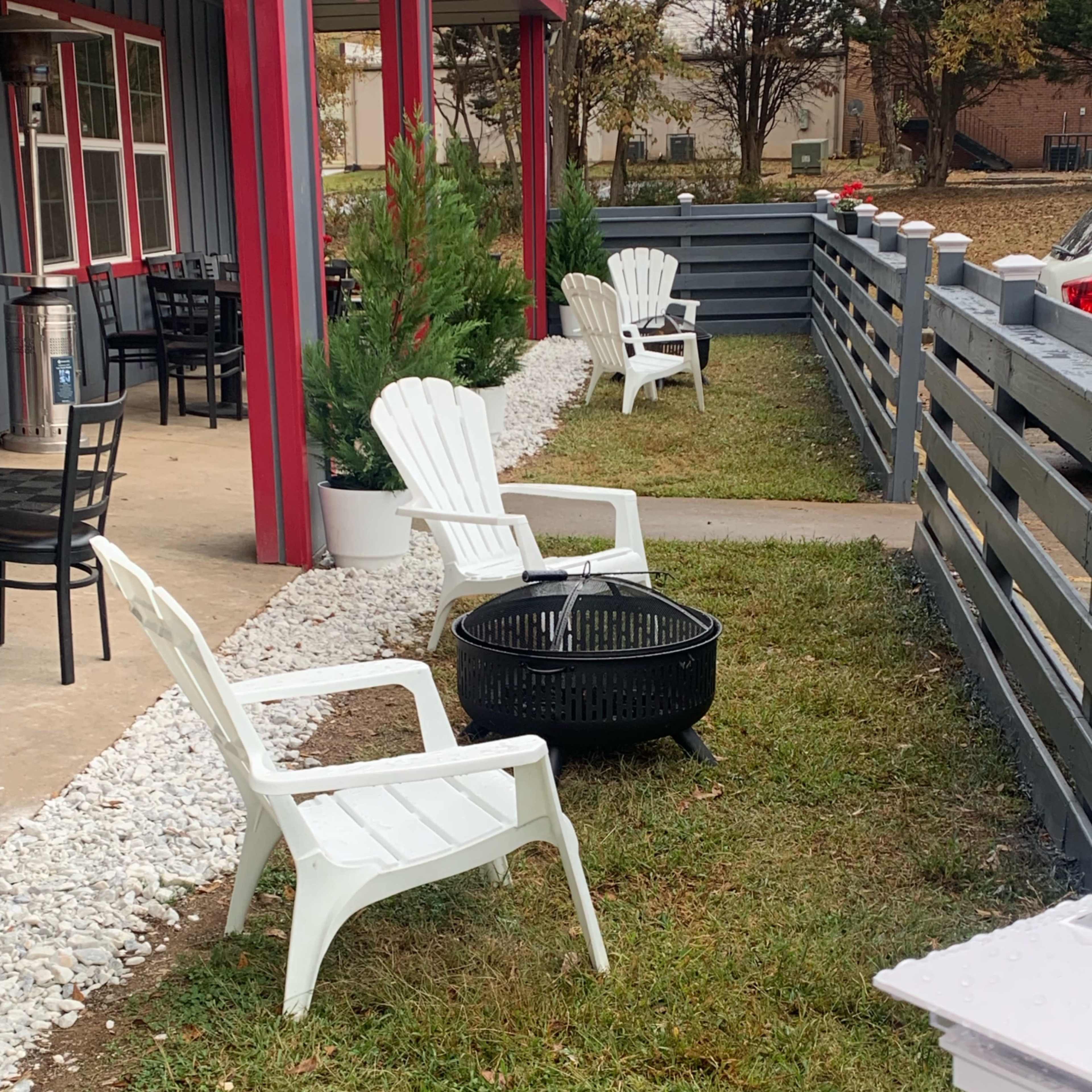 A small outdoor seating area with white plastic chairs arranged around a circular fire pit, surrounded by grass and gravel pathways.