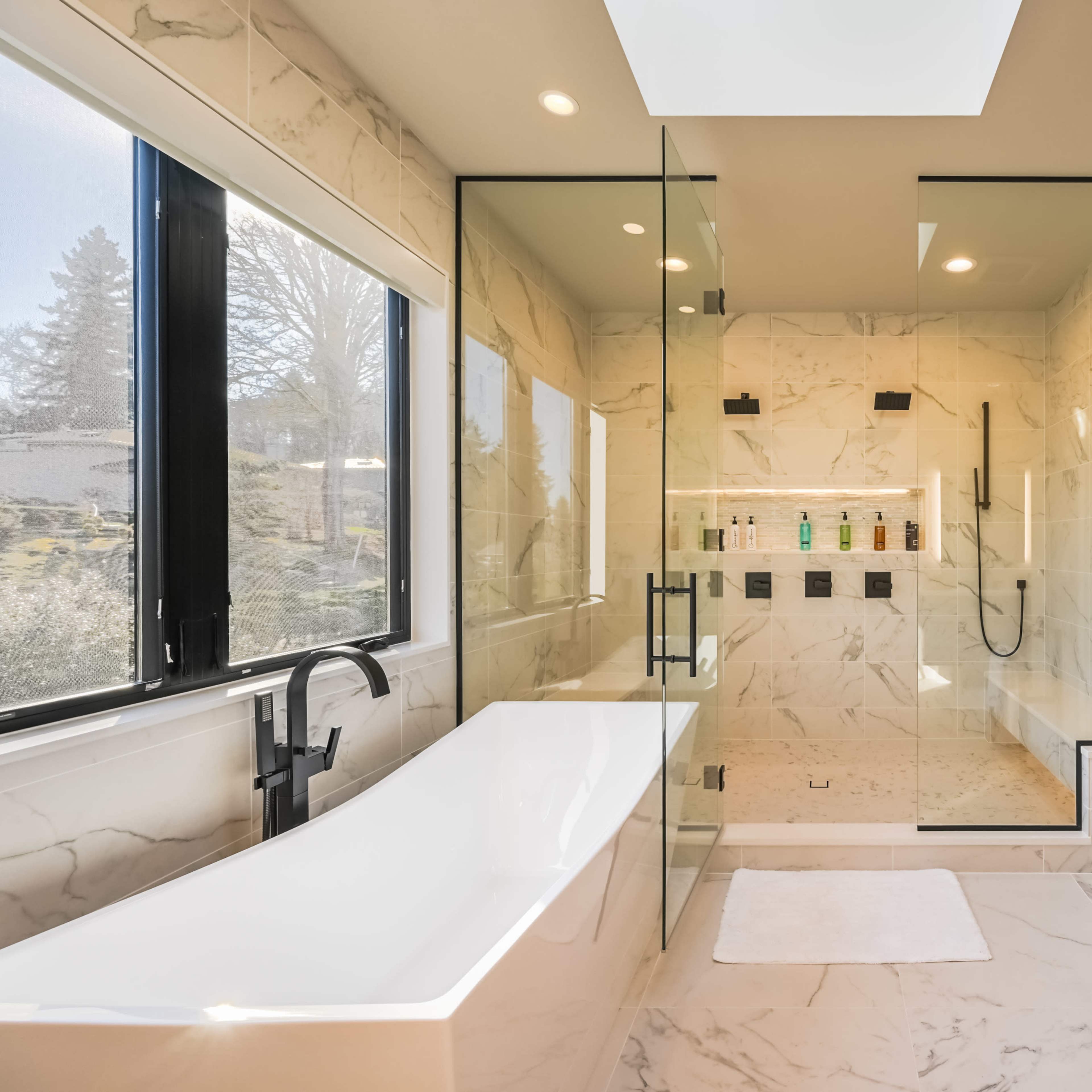 A modern bathroom featuring a glass-enclosed shower, a freestanding bathtub, and a double vanity with natural light streaming in through a large window.