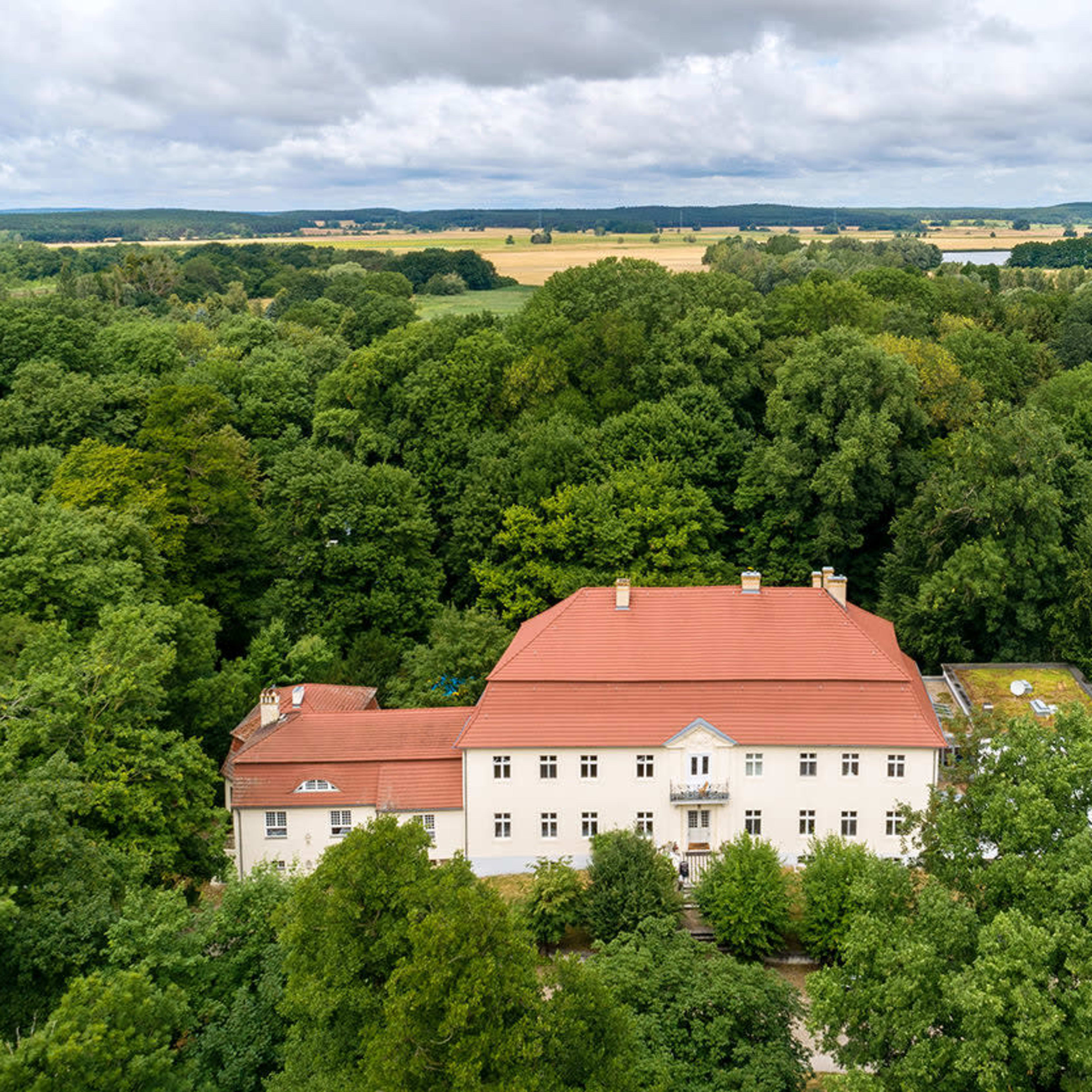 A large white building with a red roof is situated amidst dense green trees.