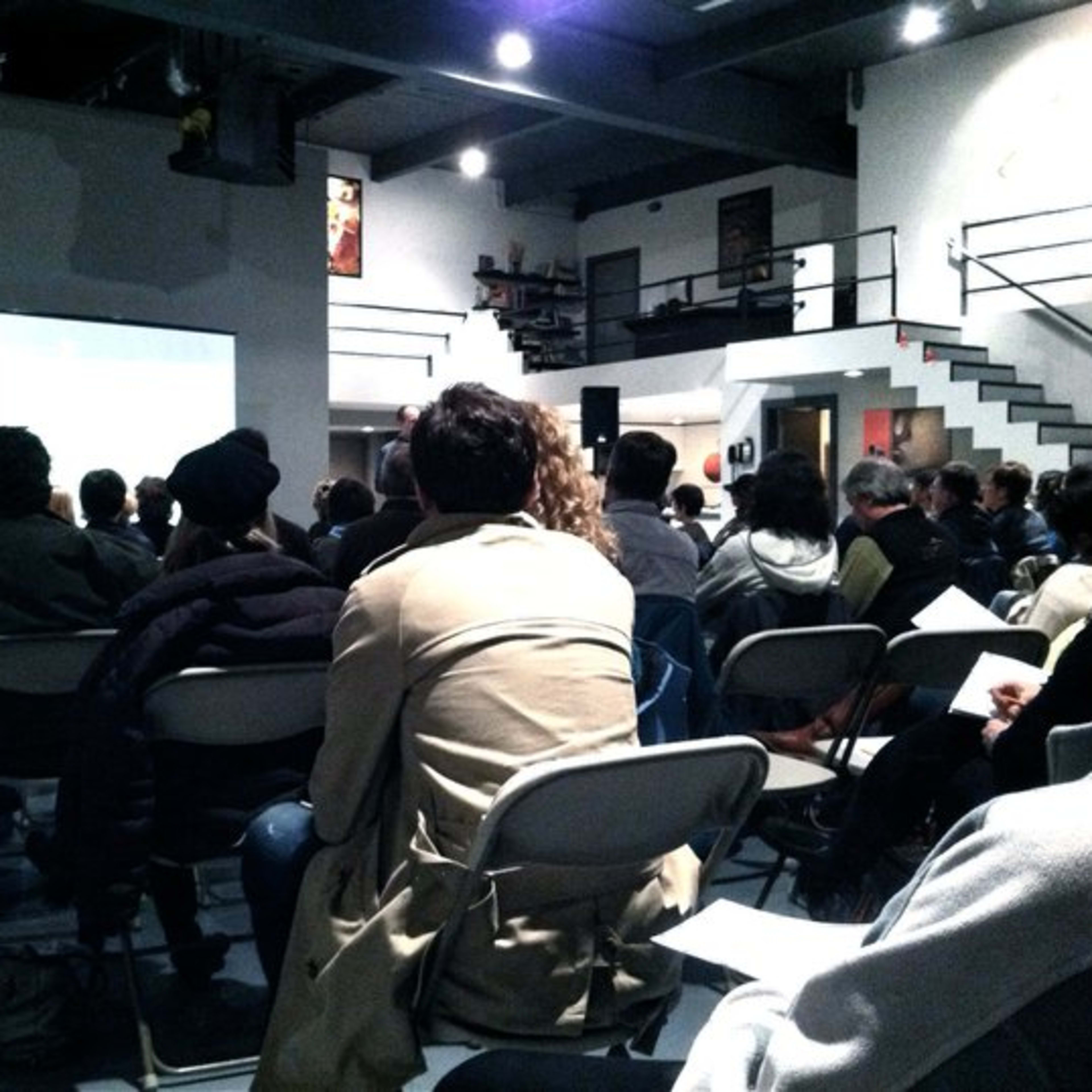 A group of people is seated in a spacious room, watching a presentation on a screen.