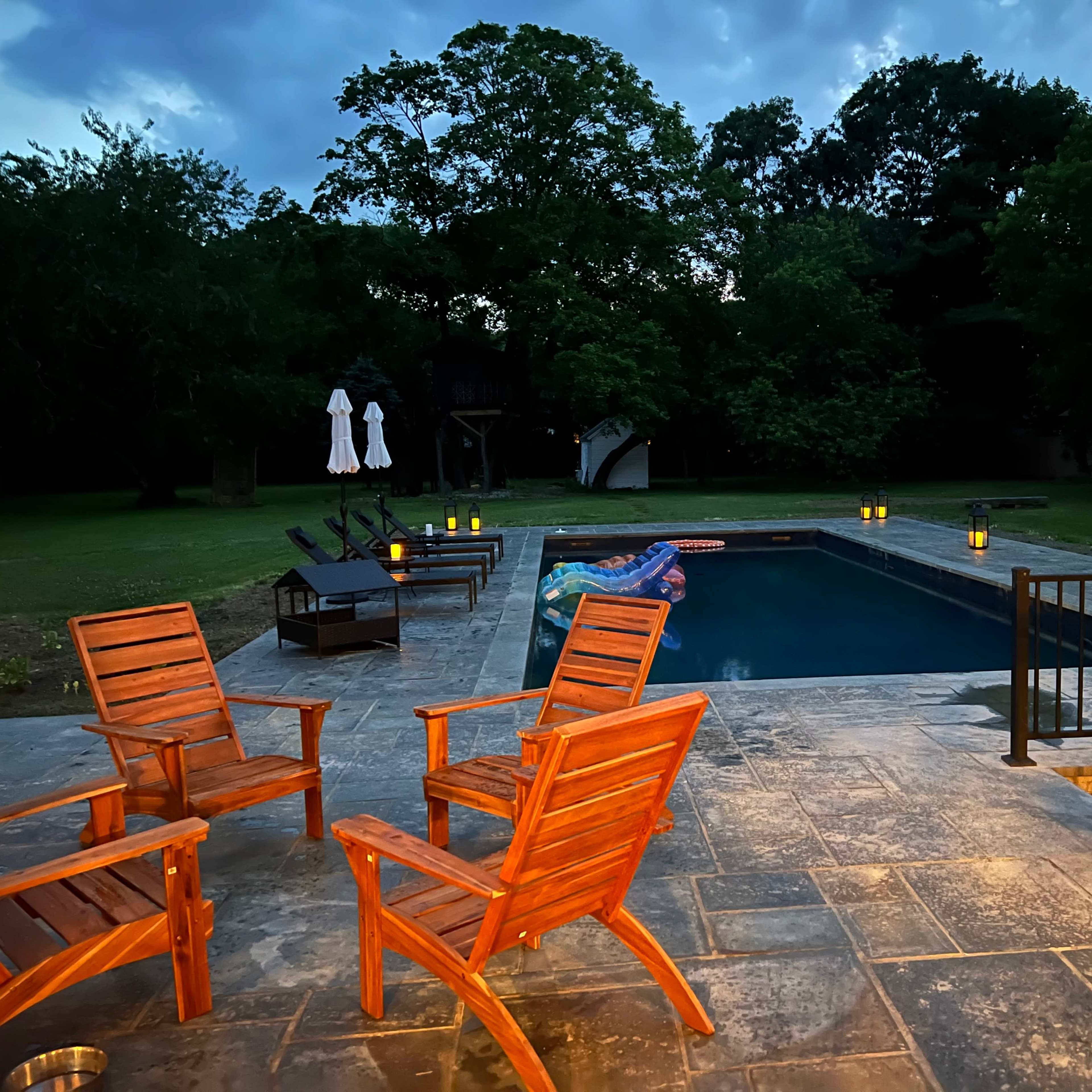 A backyard with a swimming pool surrounded by wooden chairs and lanterns, illuminated during the evening.