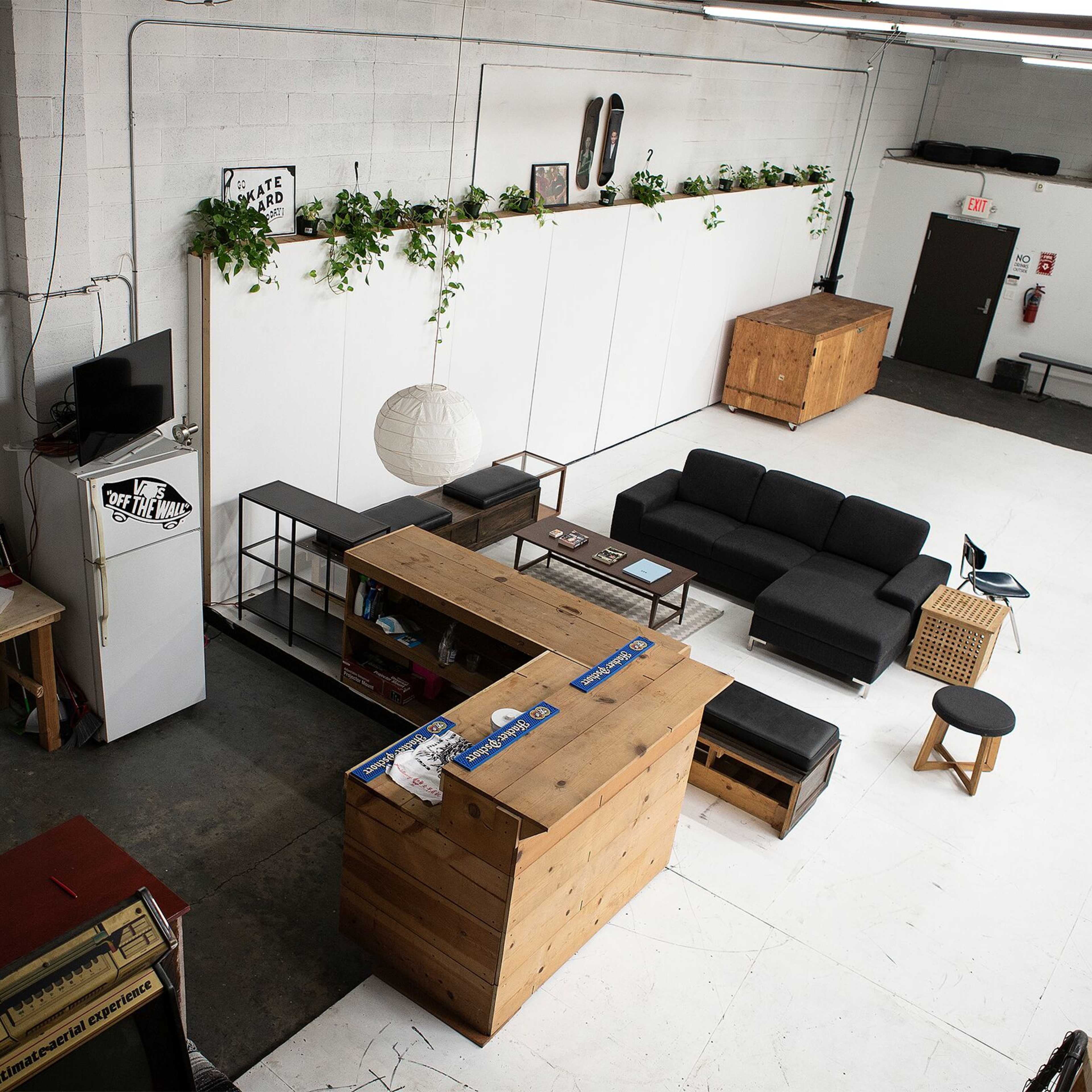 A spacious, minimalist interior with a black sofa, wooden furniture, a kitchenette area, and plants along the walls.