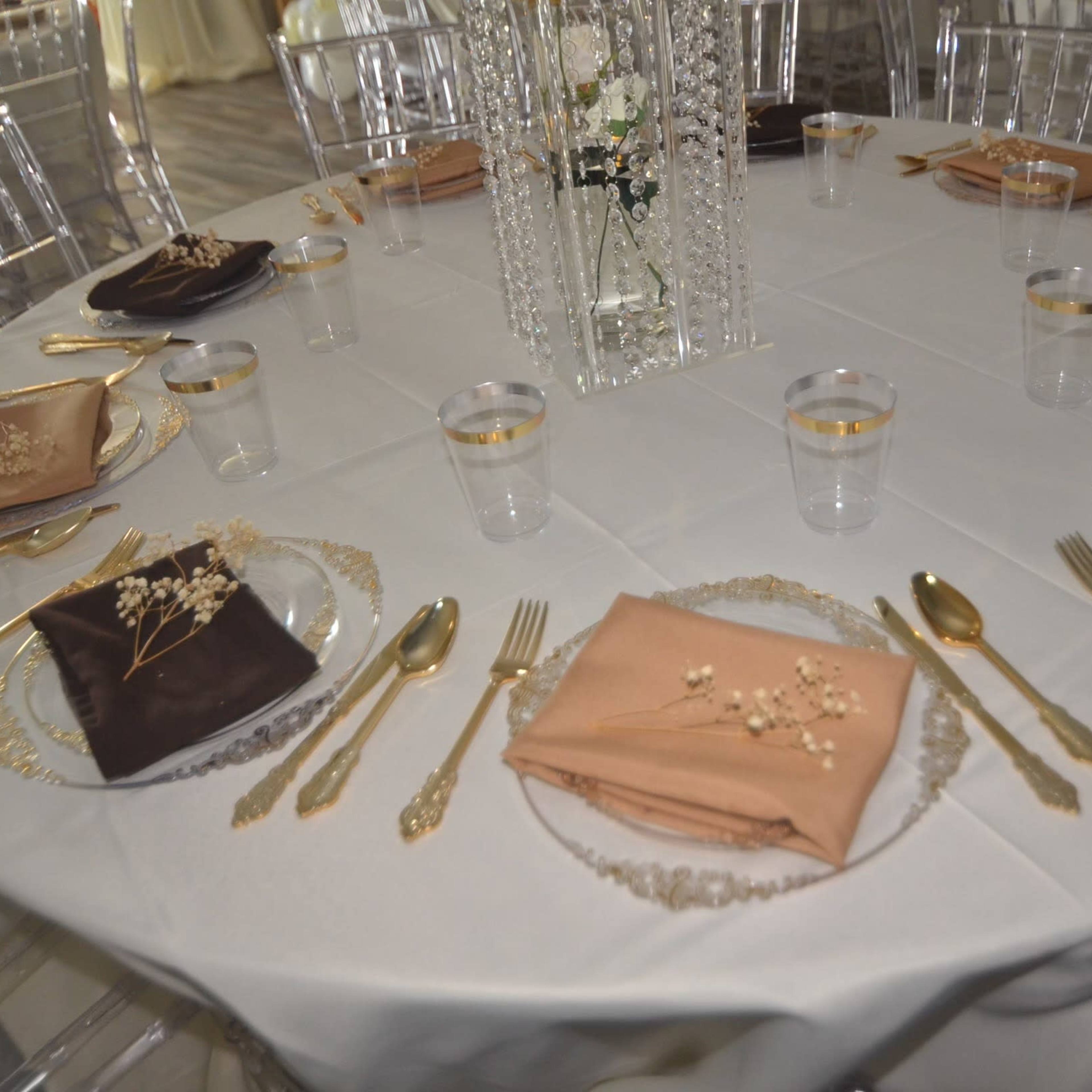 A round table is set with various plates, gold utensils, water glasses, and neatly folded napkins, with a decorative centerpiece in the middle.