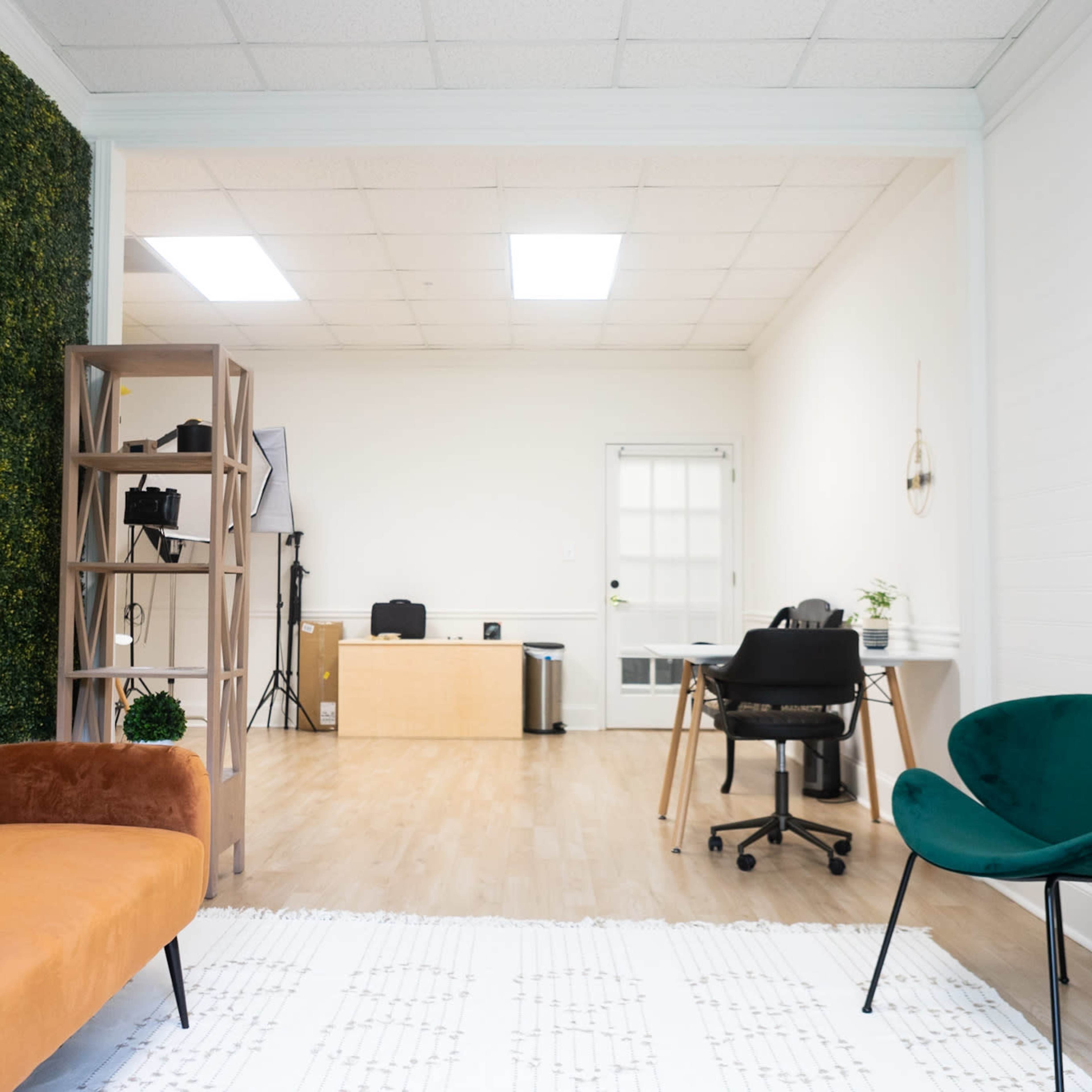 A modern office space with a green wall, an orange sofa, a wooden shelf, and a workspace featuring a desk and a black chair.