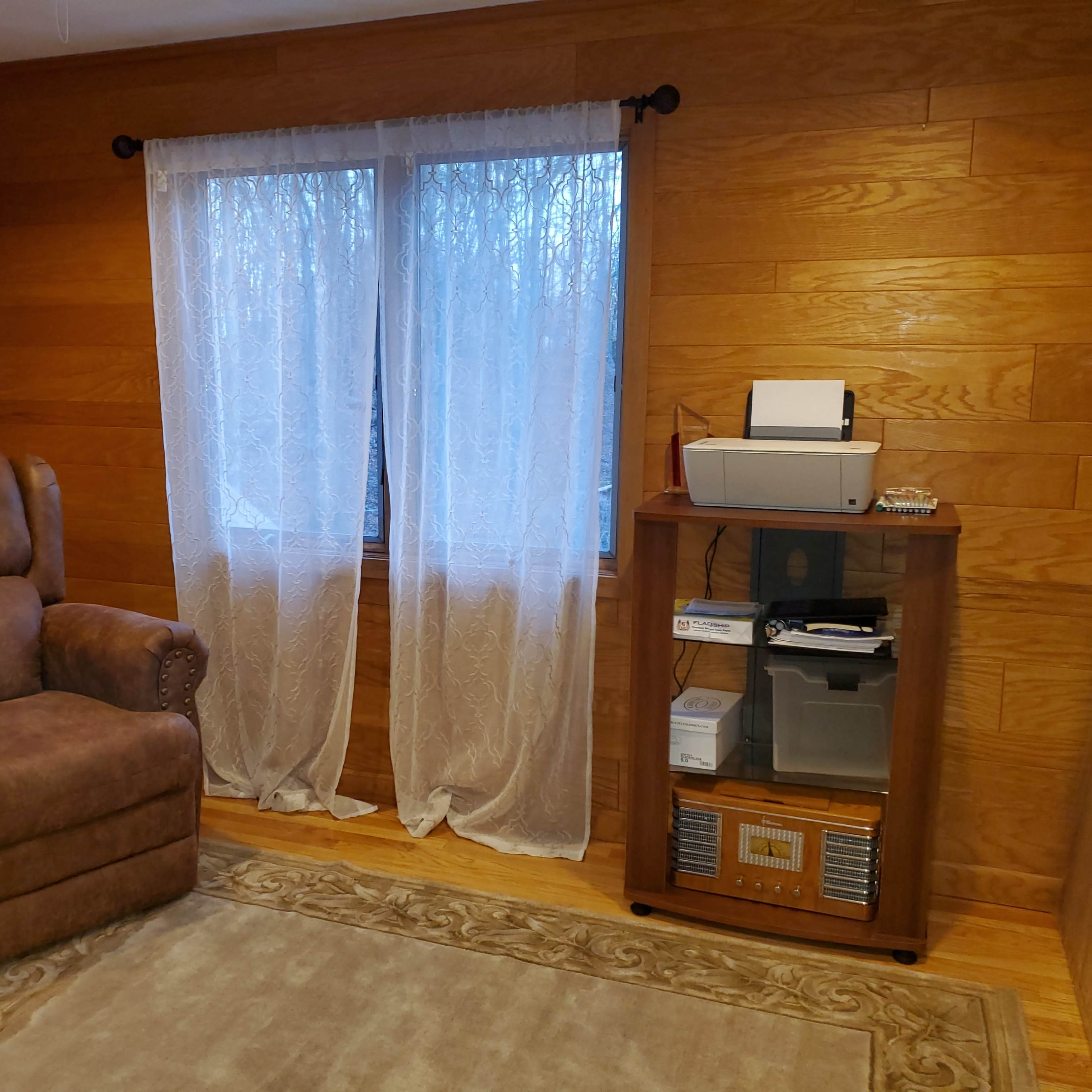 The room features a brown recliner chair beside a wooden cabinet containing a printer and various office supplies, with sheer curtains covering the window.