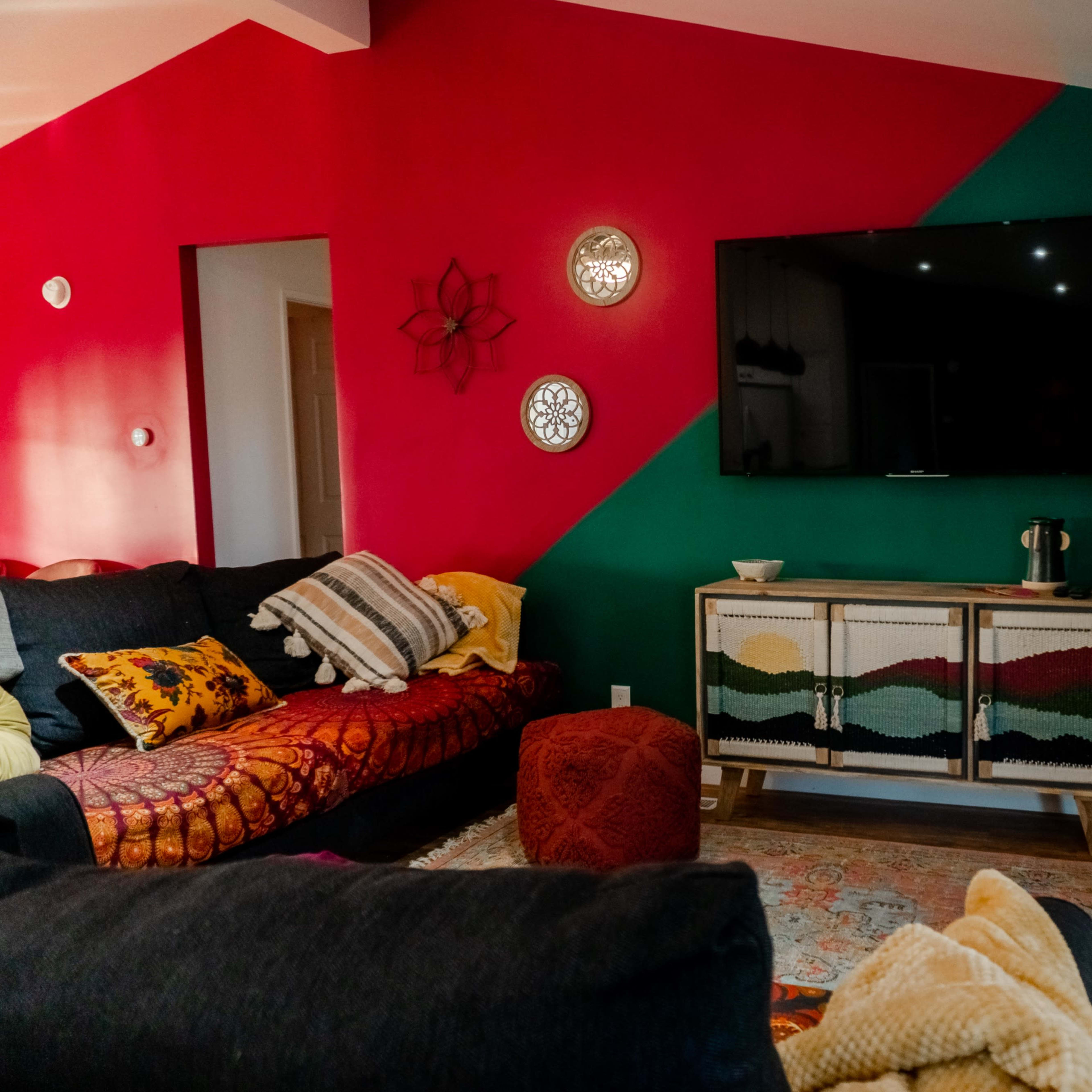 A living room features a dark sofa, colorful accent walls, a television mounted above a wooden cabinet, and decorative items like a rug and plants.