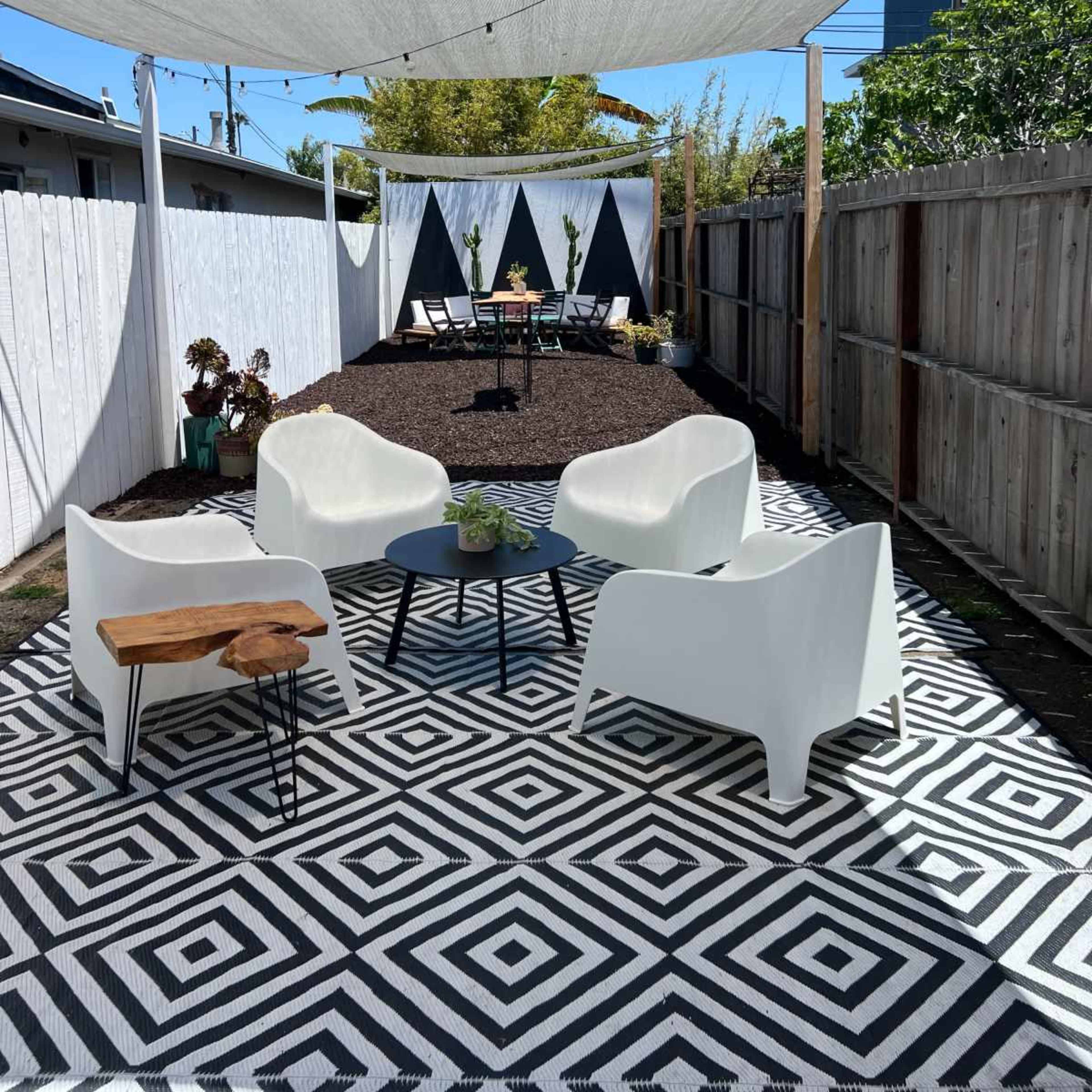A backyard space features a patterned black and white rug, modern white chairs, a small wooden table, and a shaded area with a triangular backdrop.