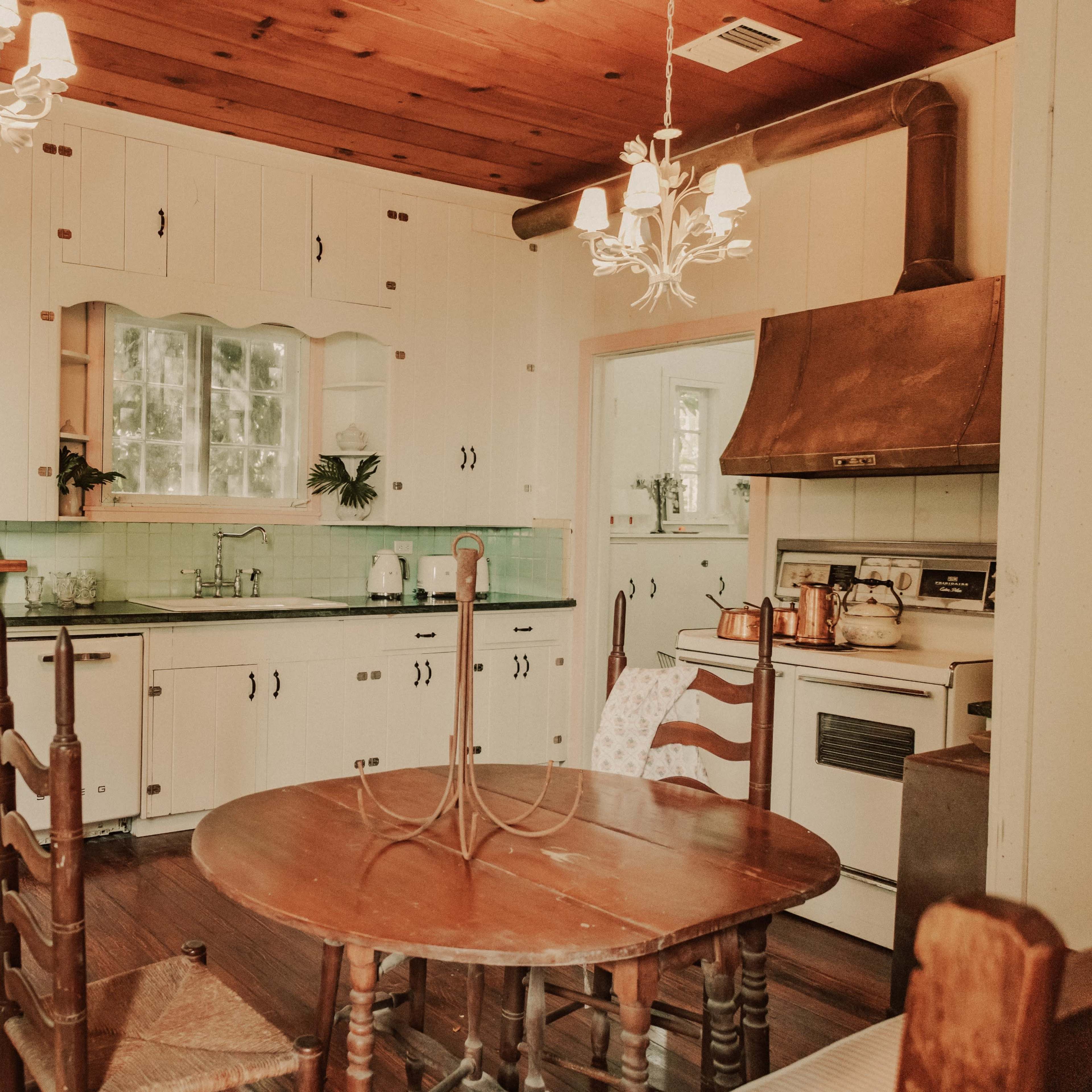 A vintage-style kitchen with white cabinets, a wooden table and chairs, and a retro refrigerator, featuring green tile accents and a stove with a chimney.