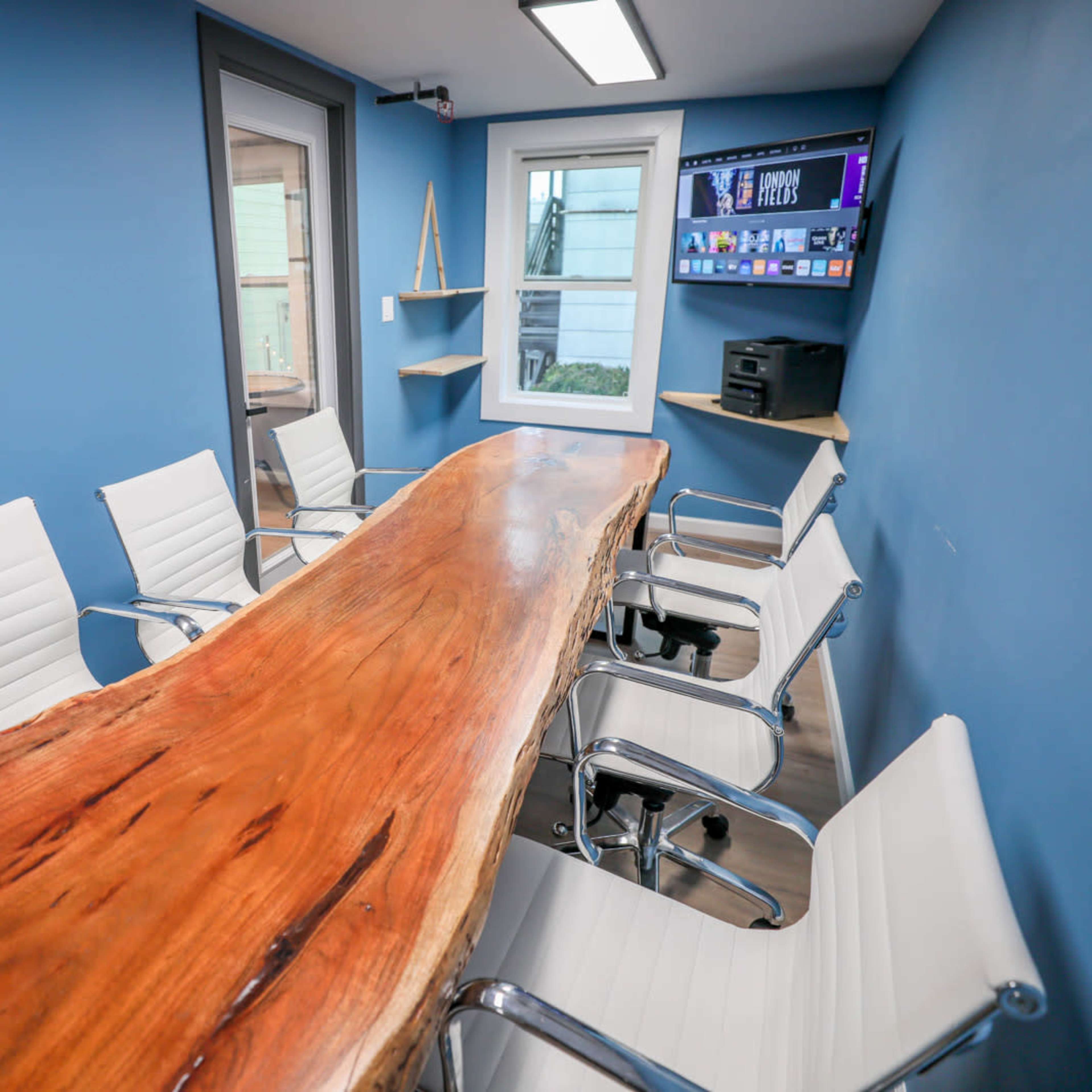 A modern conference room features a large wooden table surrounded by white chairs, with a blue wall and a window.