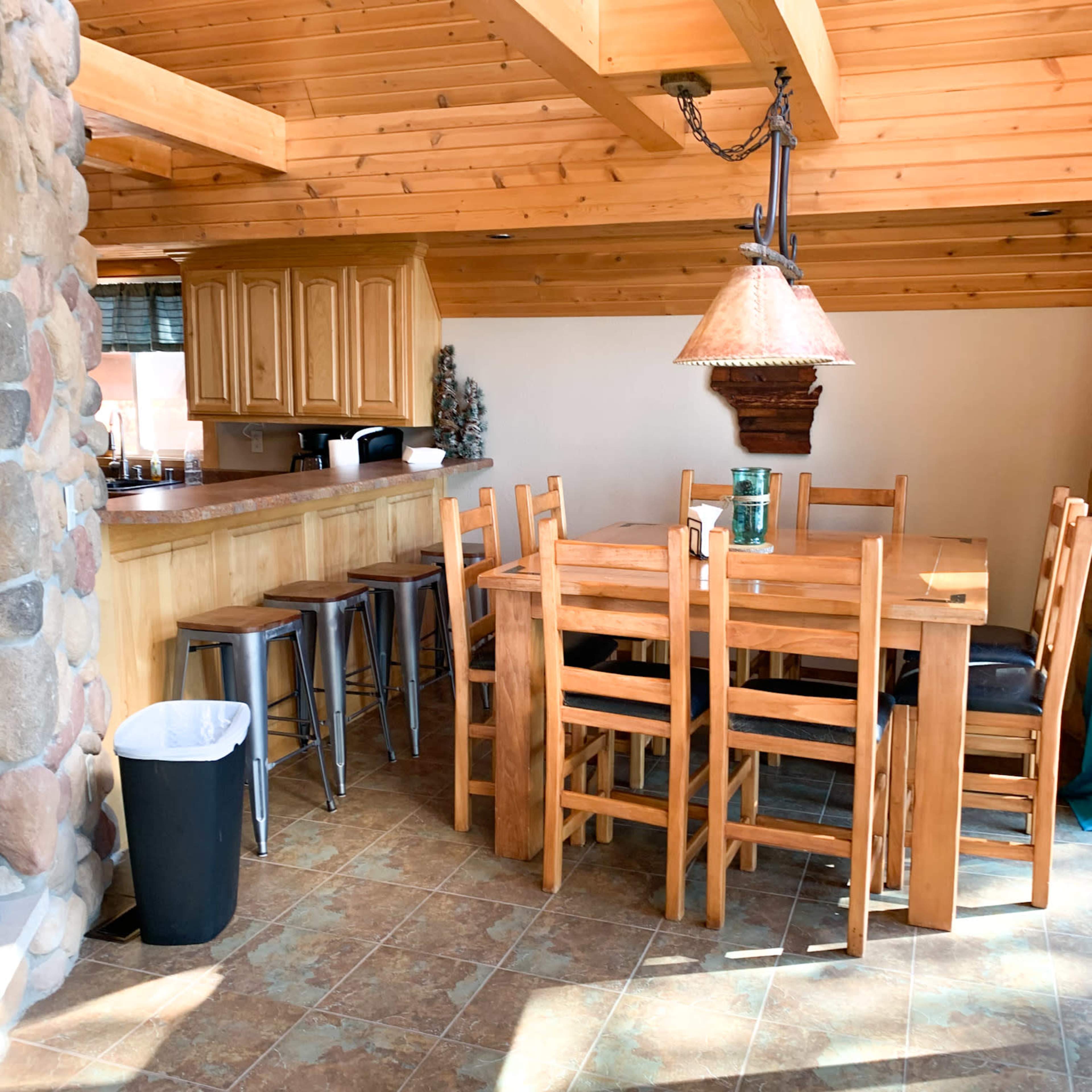 A wooden dining area with a large table surrounded by chairs, next to a kitchen with cabinetry and a counter, set against a backdrop of stone and wooden beams.