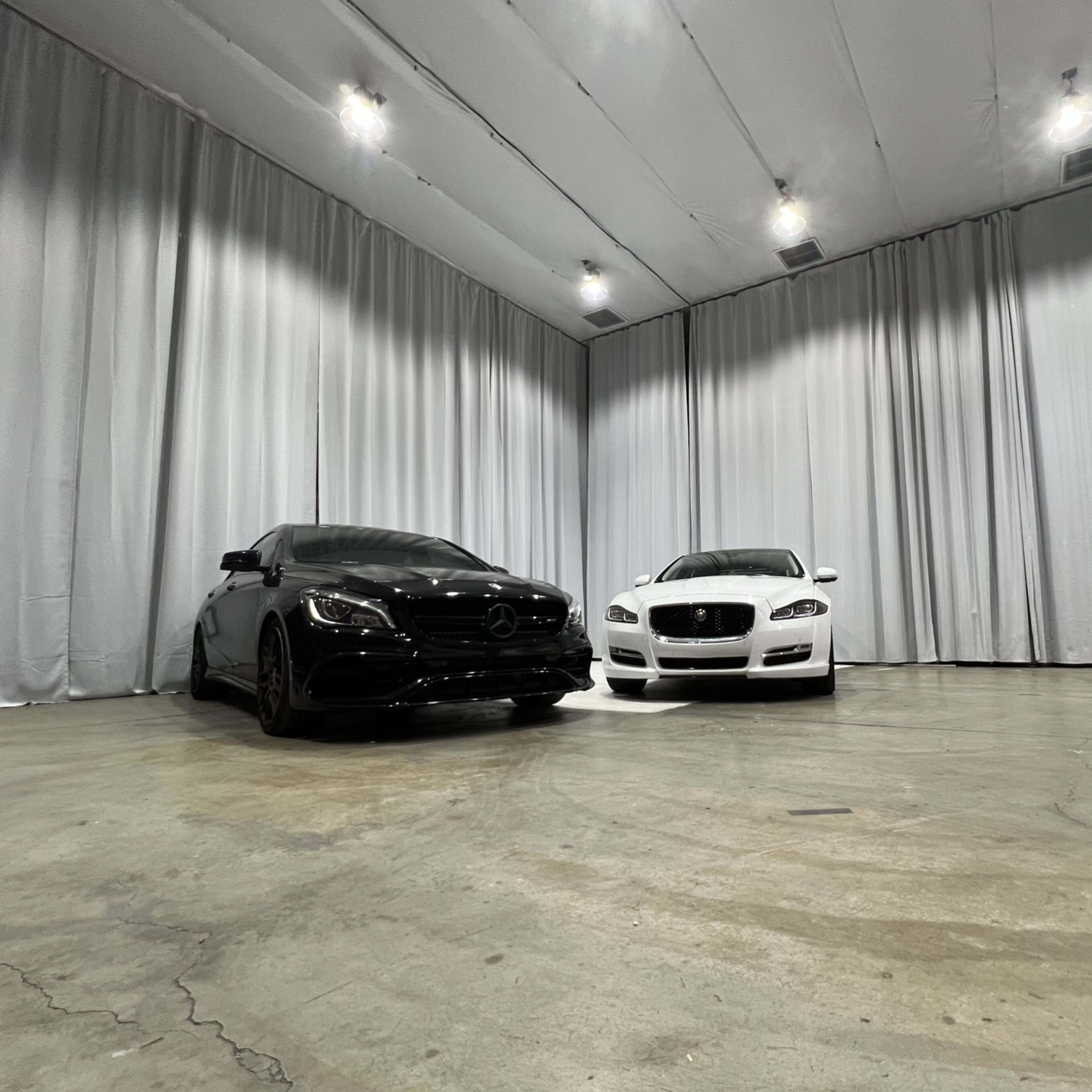 A black Mercedes-Benz and a white Jaguar are parked side by side in a spacious, minimalist room with white draped curtains.
