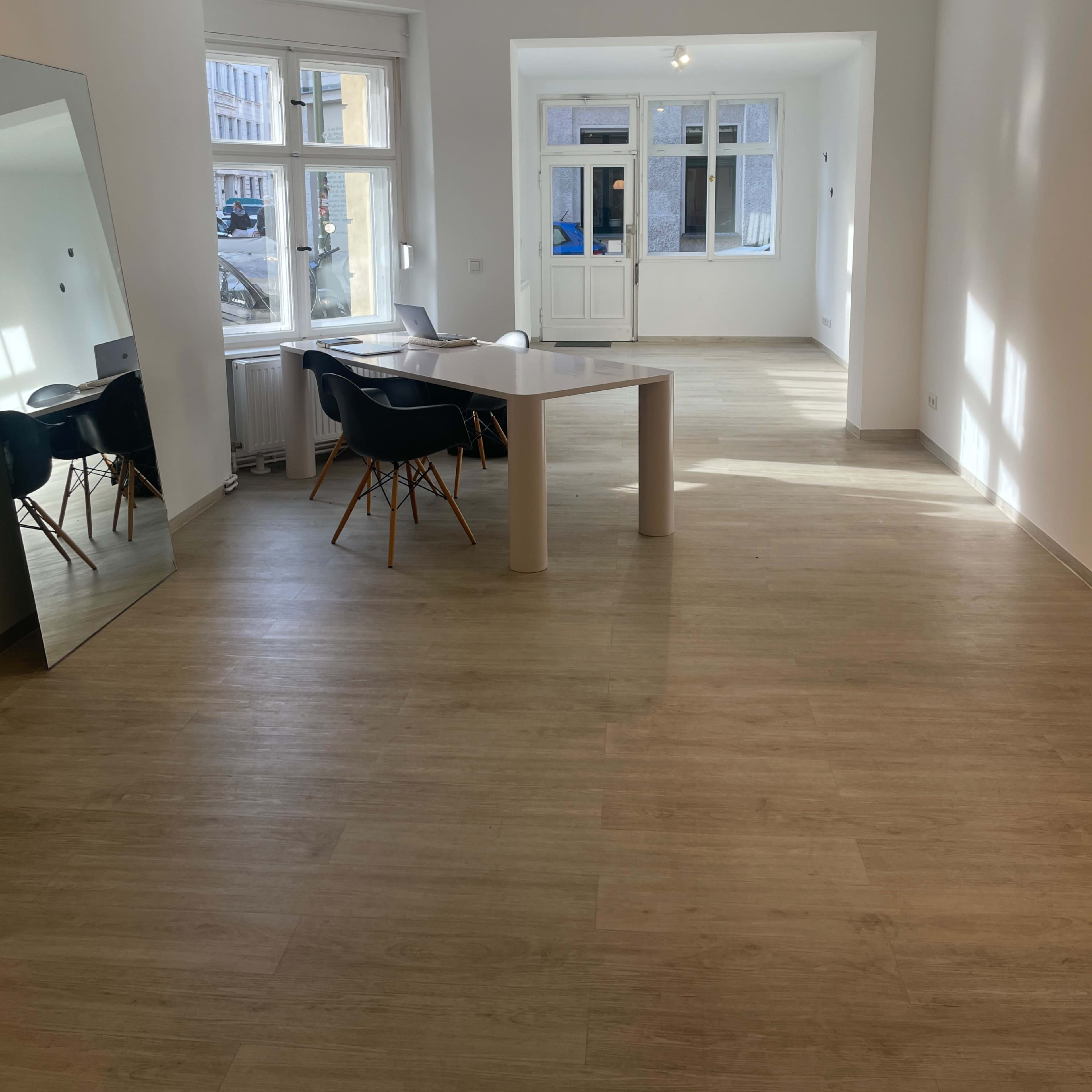 A spacious, empty room with light-colored walls, a wooden floor, a table with chairs, and large windows allowing natural light to enter.