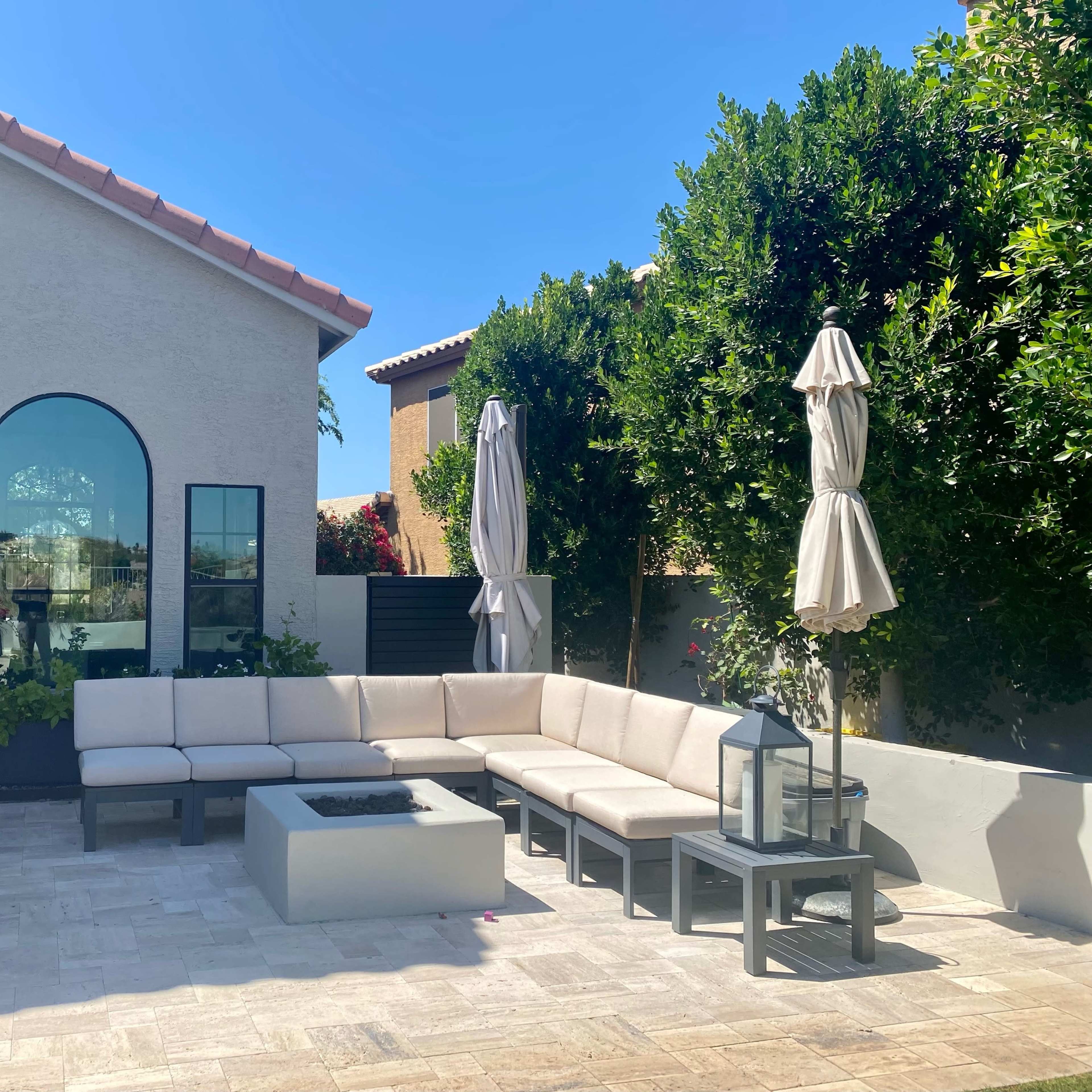 A shaded outdoor seating area with a modern sectional couch, a fire pit, and patio umbrellas surrounded by greenery.