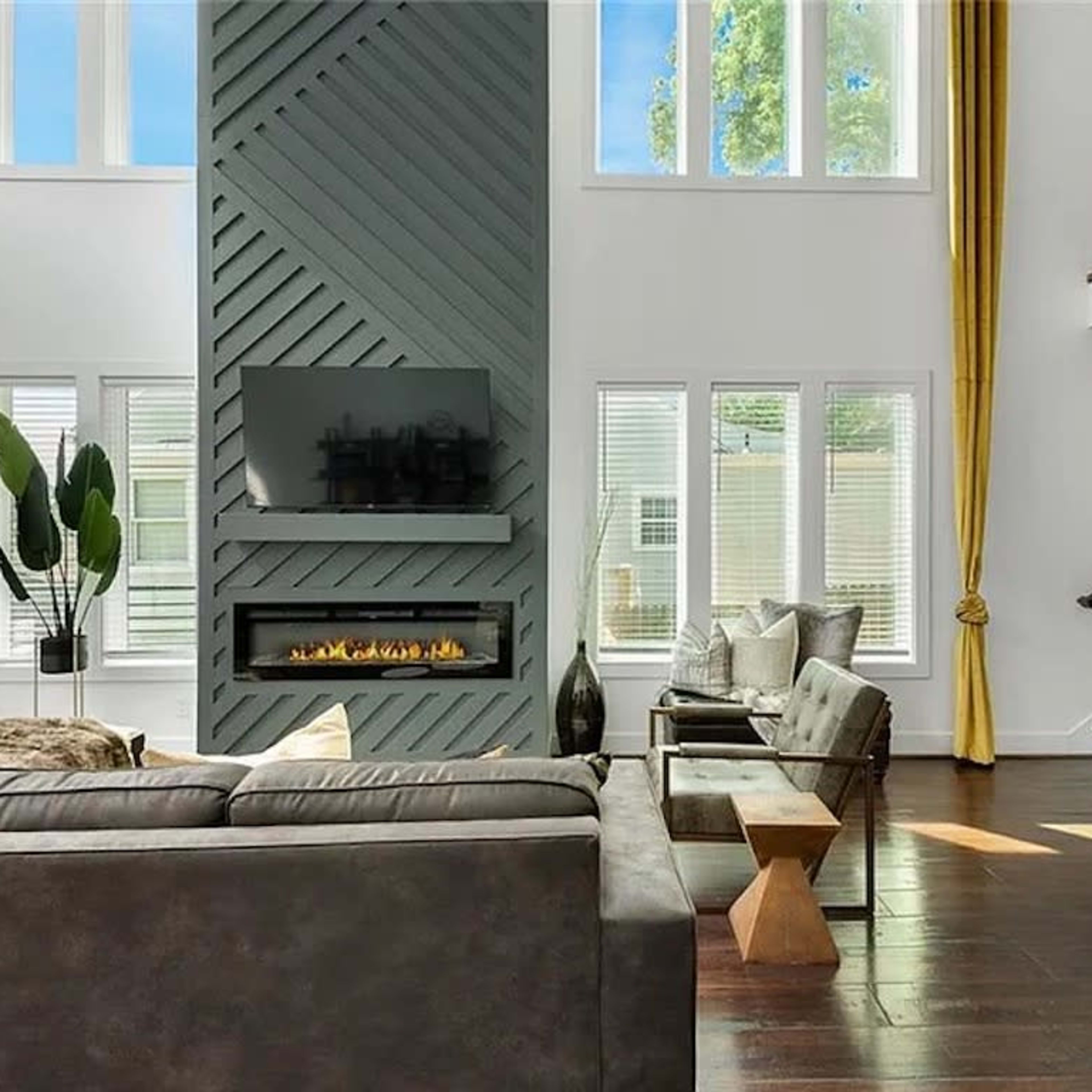 A modern living room features a tall fireplace with a TV mounted above it, surrounded by large windows and yellow curtains, alongside a sofa and minimalist furniture.