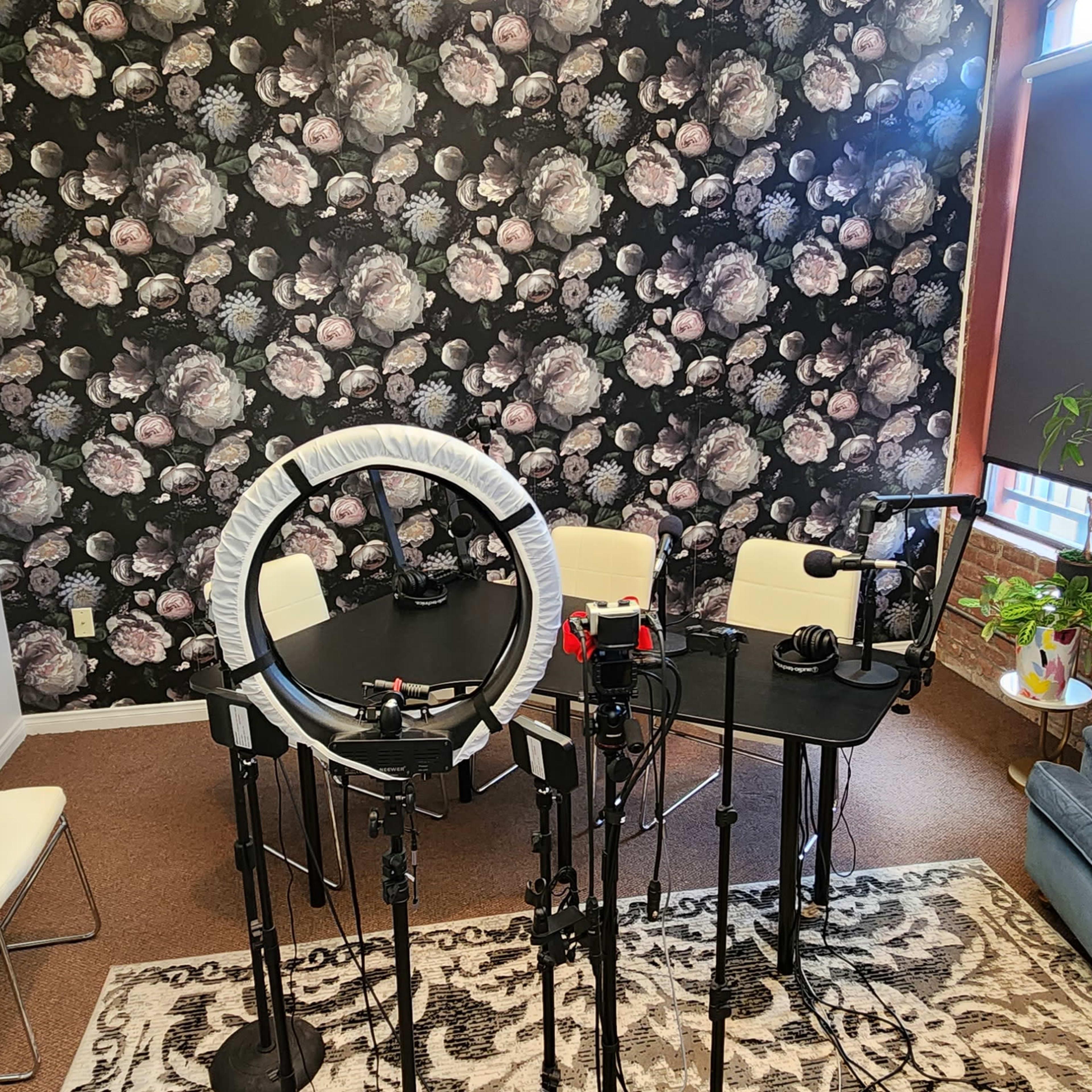 A recording studio setup with a floral wallpaper background, a table with microphones, chairs, and lighting equipment.