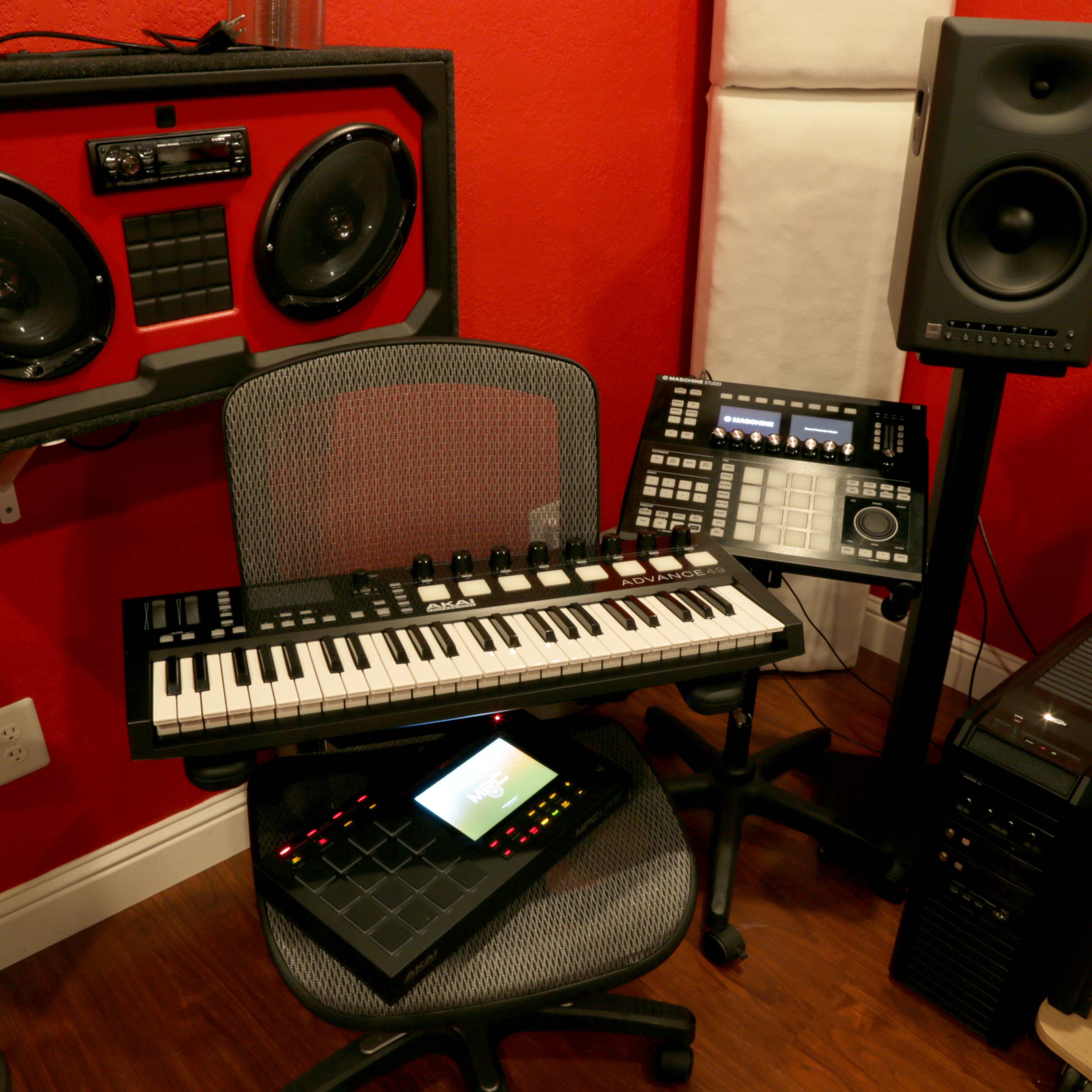 A music production workspace featuring a keyboard, a drum pad, and studio monitors against a red wall.