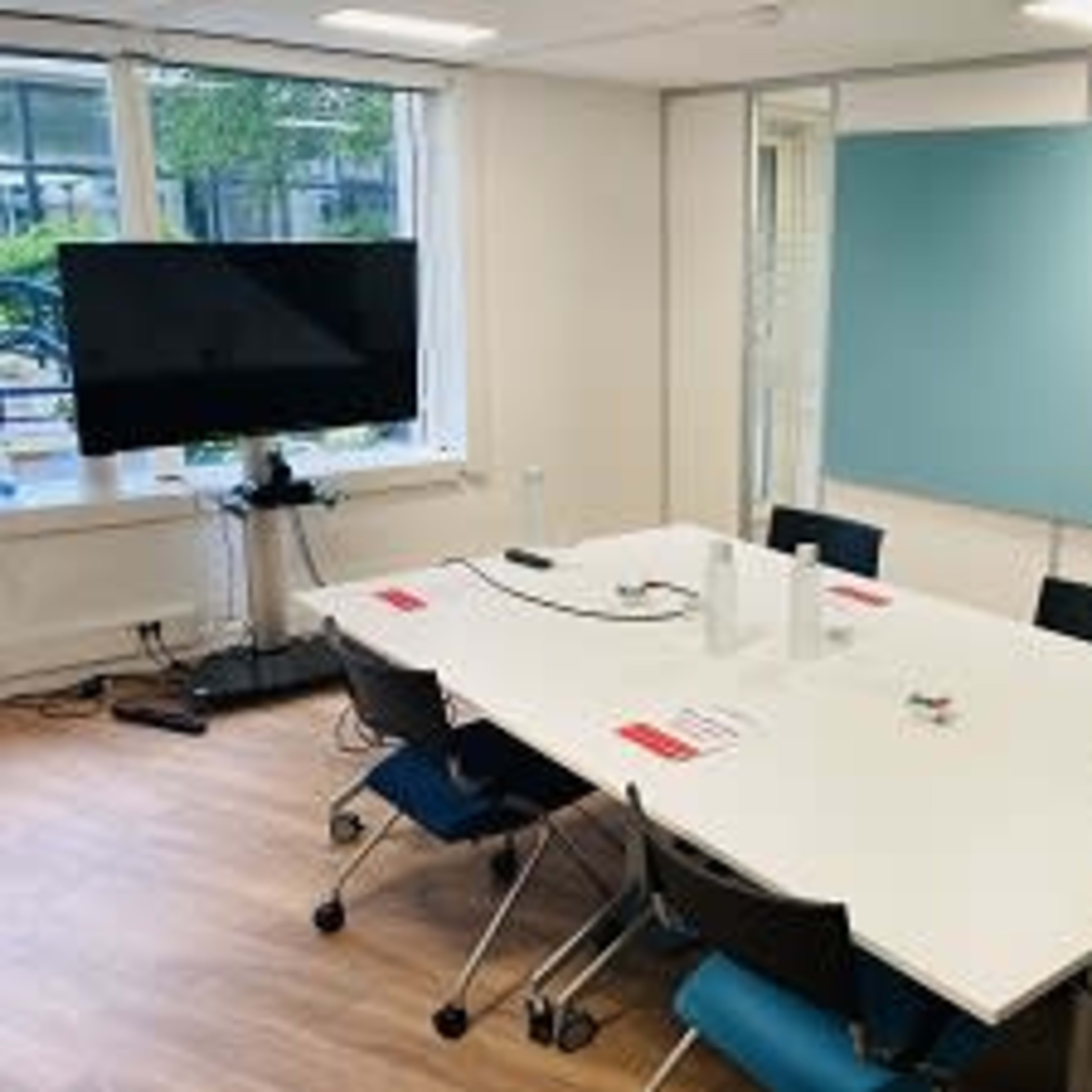 A meeting room features a large white table surrounded by chairs, equipped with a television on a stand and a blue wall area for presentations.