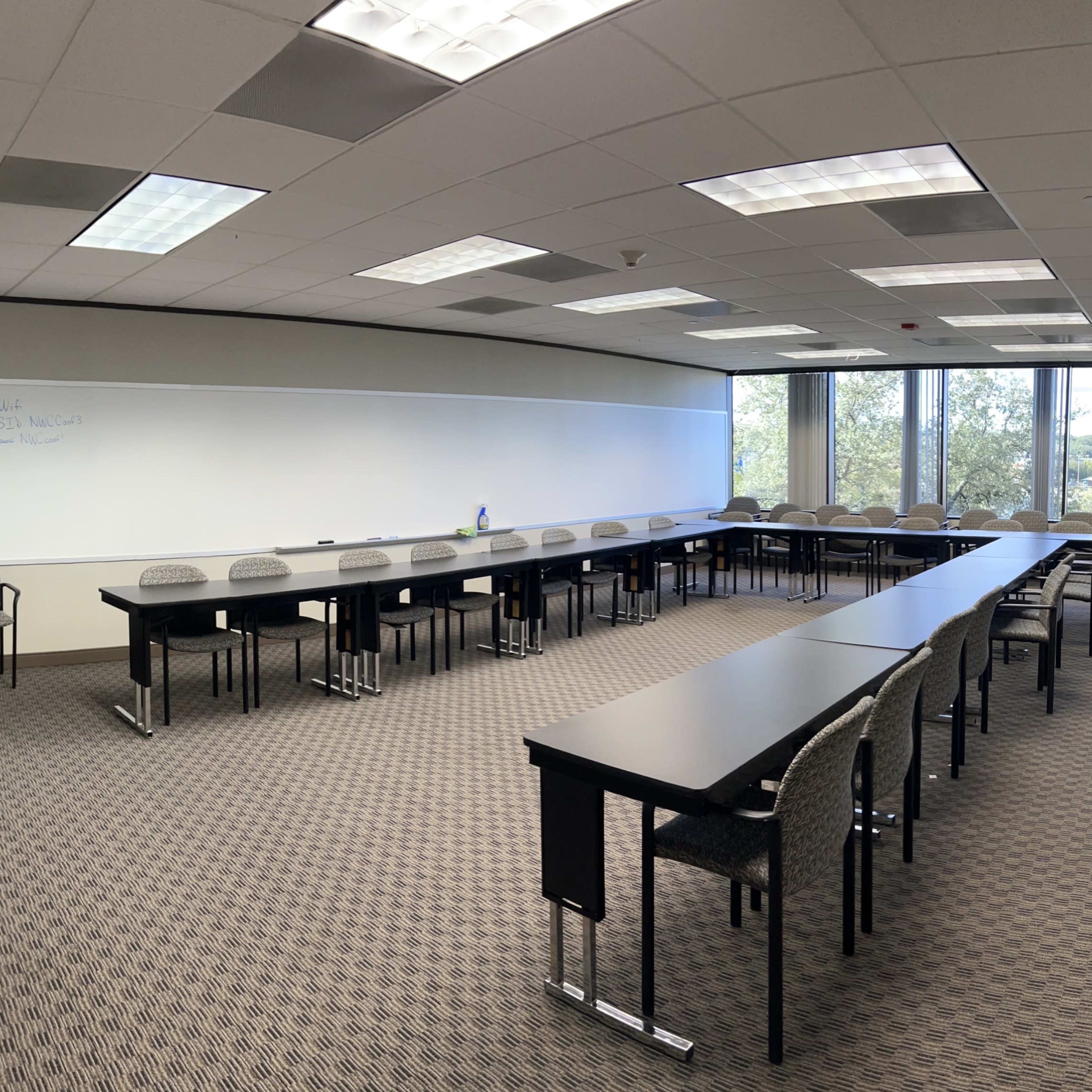 A large conference room features multiple long tables arranged in a U-shape with chairs, illuminated by natural light from windows on one side.