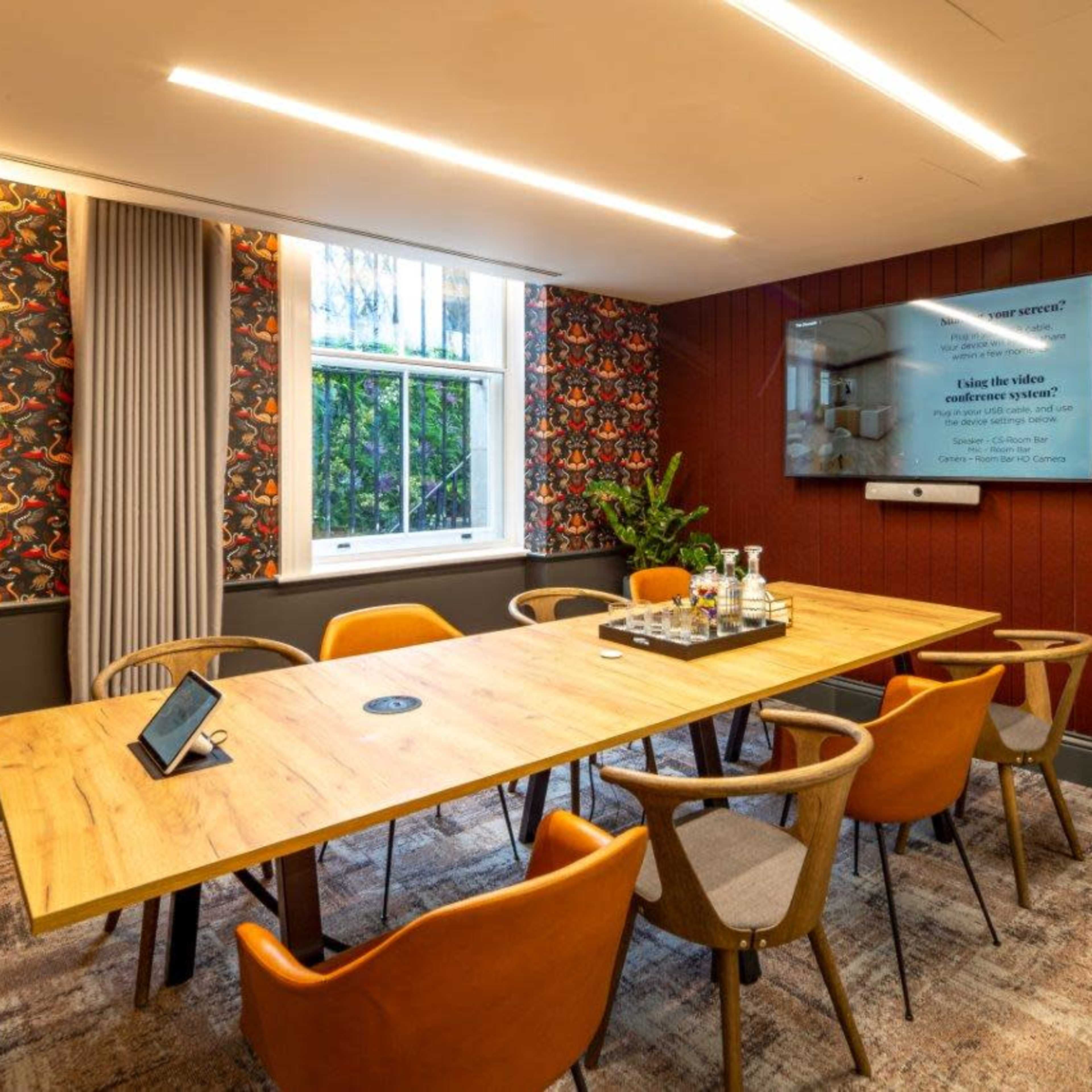A modern meeting room featuring a long wooden table surrounded by six chairs, potted plants, a large screen on the wall, and decorative wallpaper.